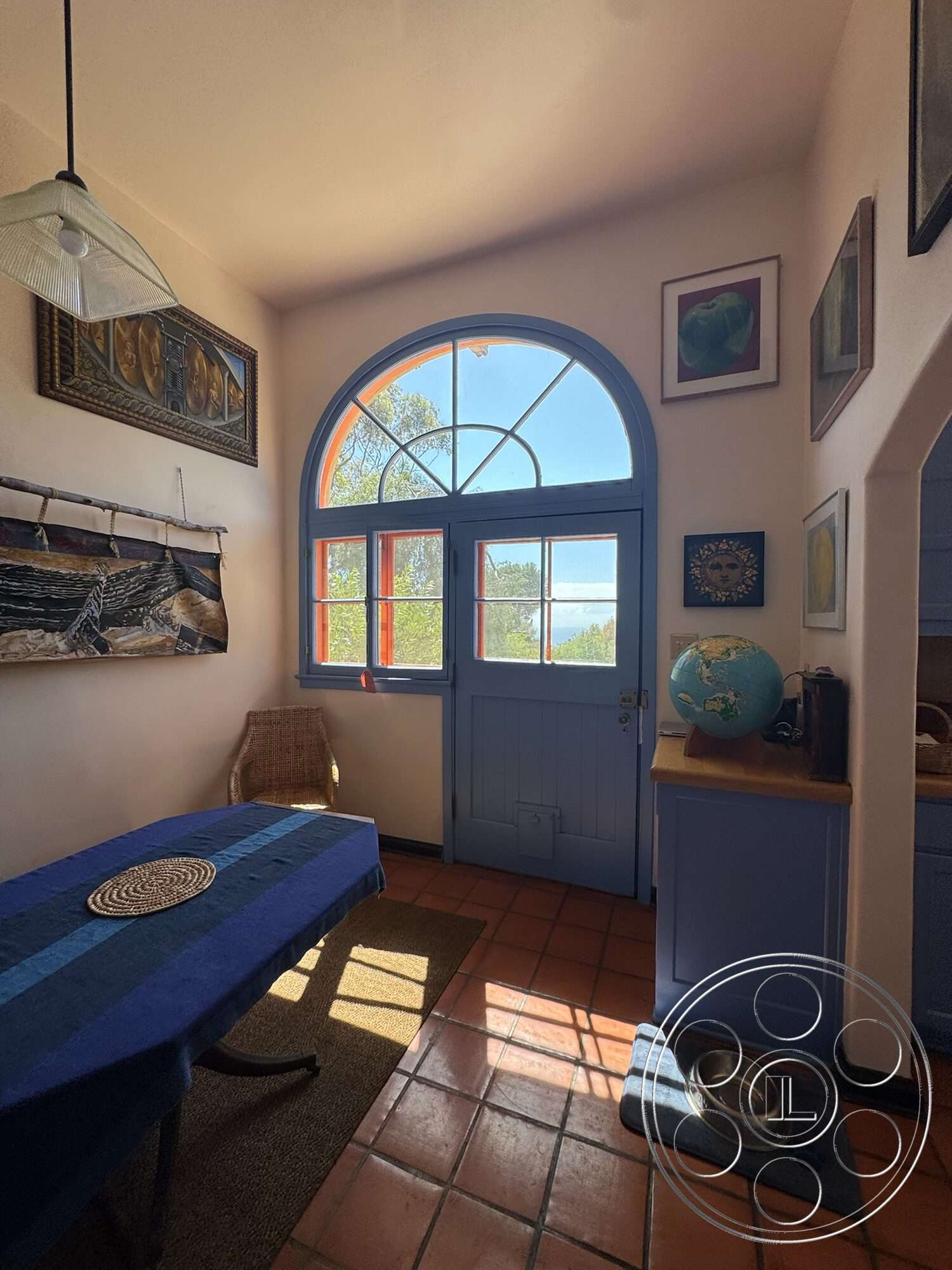 Farmstead Garden - wooden dining table, framed artwork, arched entryway, textured wall treatment, glass pendant light, decorative globe, wicker chair, woven table runner, blue painted wooden door, terra-cotta floor tiles, blue kitchen cabinets