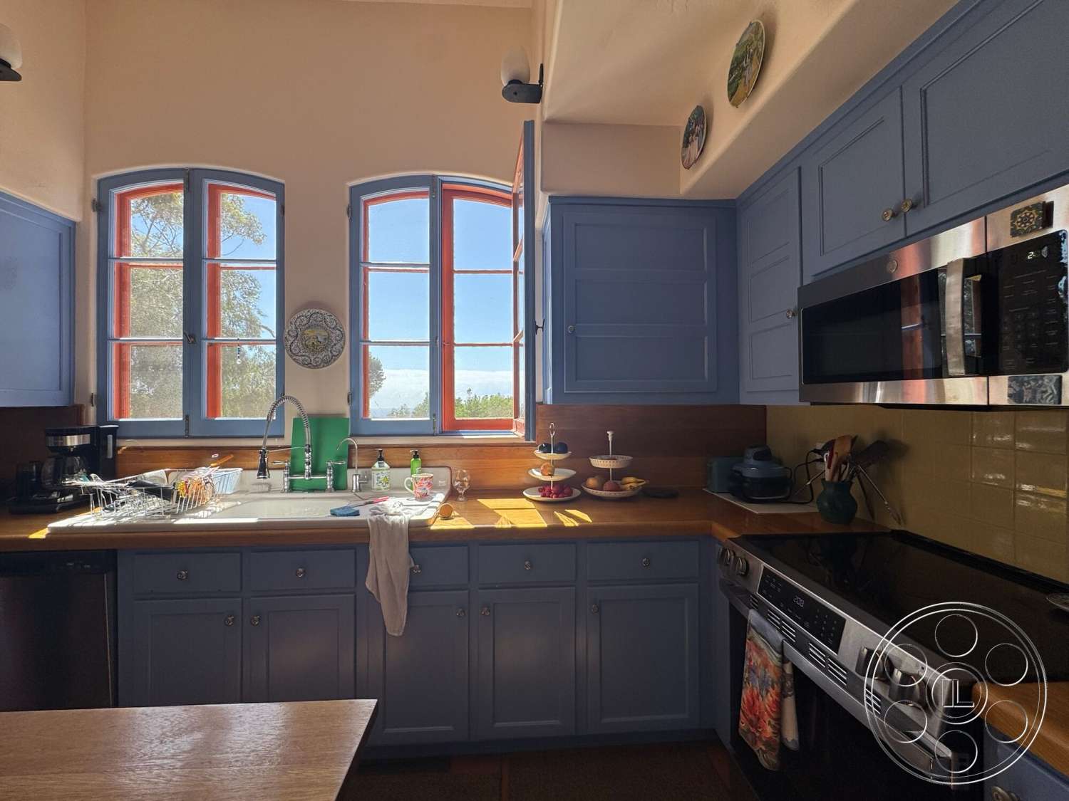 Farmstead Garden - natural light, stainless steel appliances, open shelving, kitchen island, wooden countertop, coffe maker, ceramic tile backsplash, double-hung windows, wood beam ceiling, blue kitchen cabinets, green kitchen faucet, glass dishware, fruit stand, rustic kitchen decor
