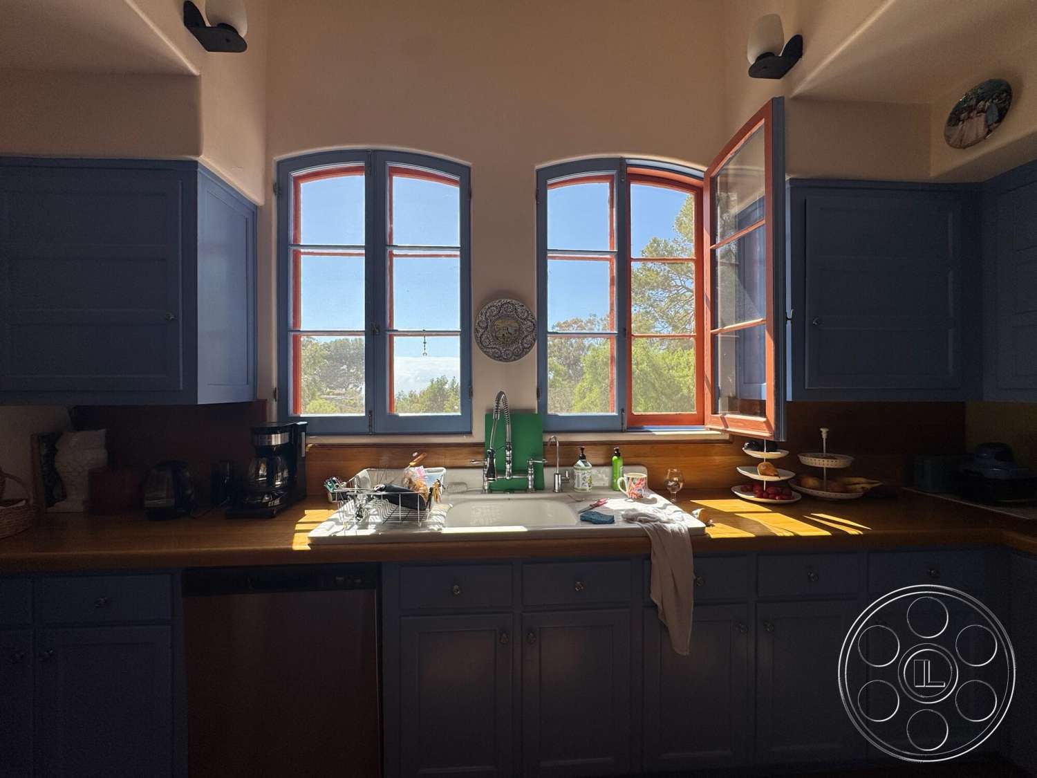 Farmstead Garden - bright natural lighting, wood countertop, kitchen sink, open window view, modern kitchen design, wall-mounted light fixtures, stainless steel dishwasher, green kitchen faucet, blue wooden cabinetry, double-hung windows with red trim, decorative wall plate