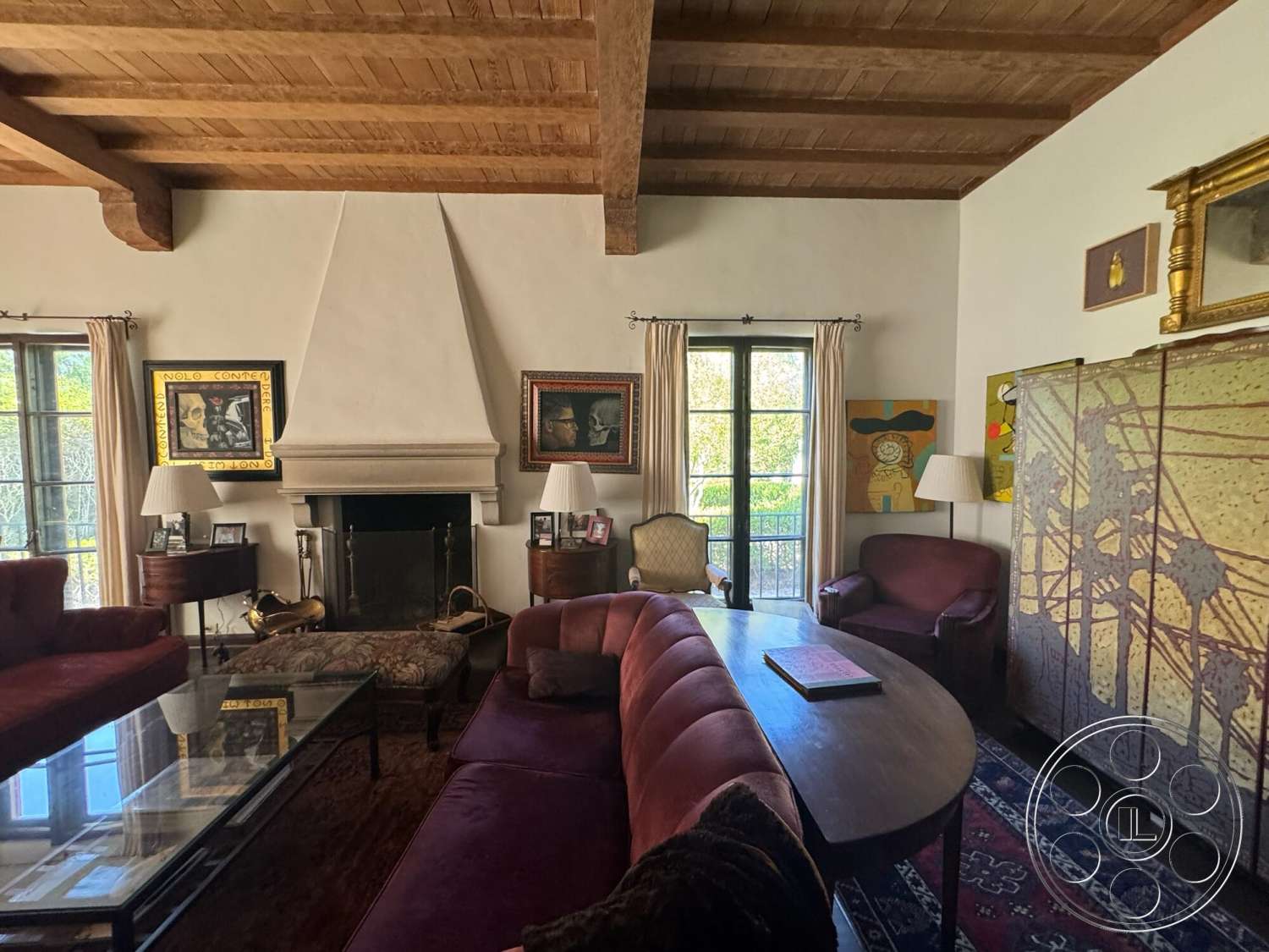 Farmstead Garden - decorative area rug, glass coffee table, floor-to-ceiling windows, framed wall art, hardwood floor, wooden side table, table lamps, plastered white walls, exposed wood ceiling beams, stone fireplace surround, vaulted living room ceiling, plush burgundy sofa, overstuffed armchairs, artistic room divider