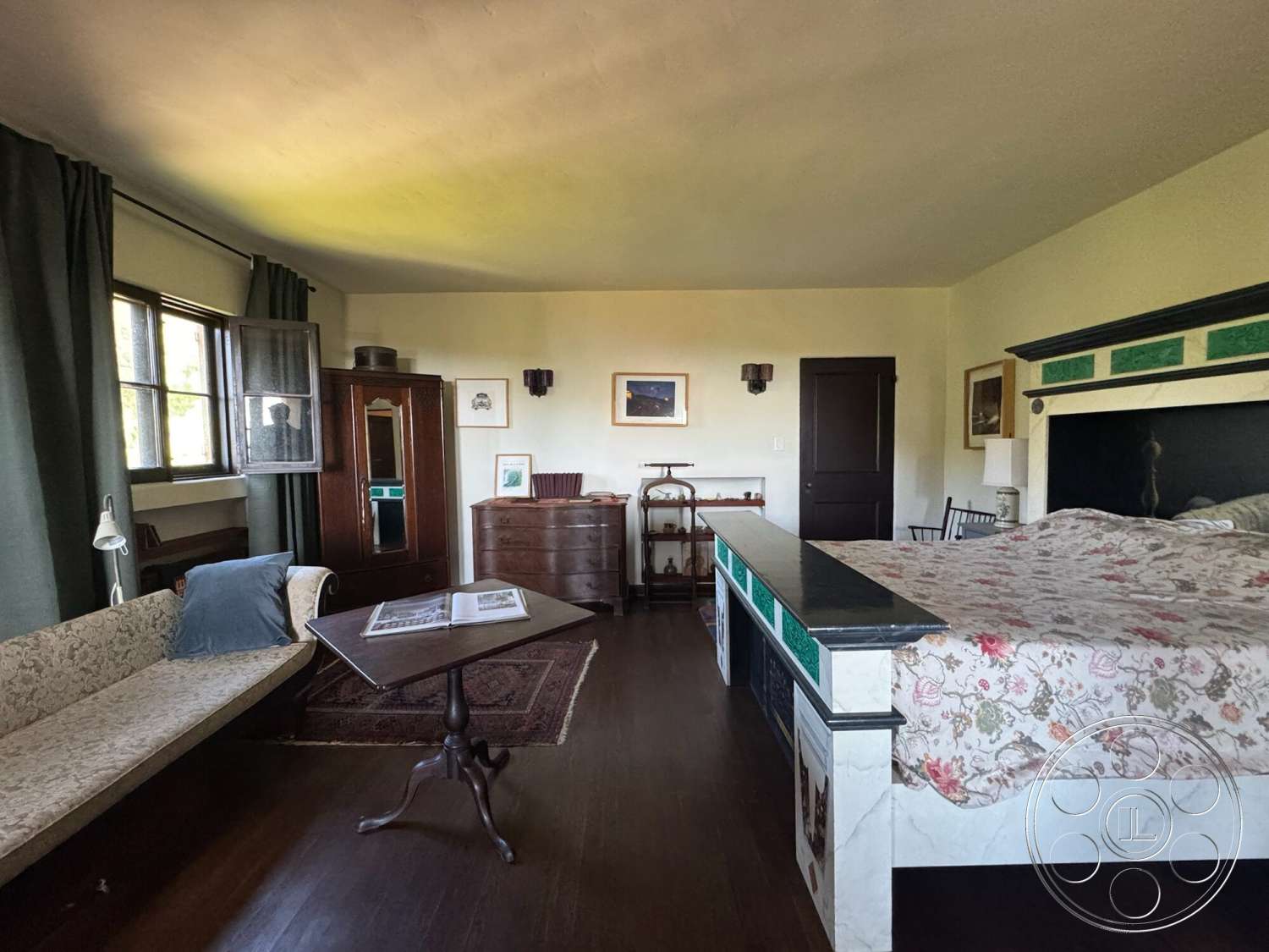 Farmstead Garden - decorative wall art, textured plaster walls, hardwood bedroom floors, large windows with dark frames, floral patterned bedding, dark wood paneling on door, vintage wooden dresser, small side table with ornate base, green fabric curtains, traditional style armchair, cabinet with glass doors, floor rug with intricate design, simple ceiling light fixture