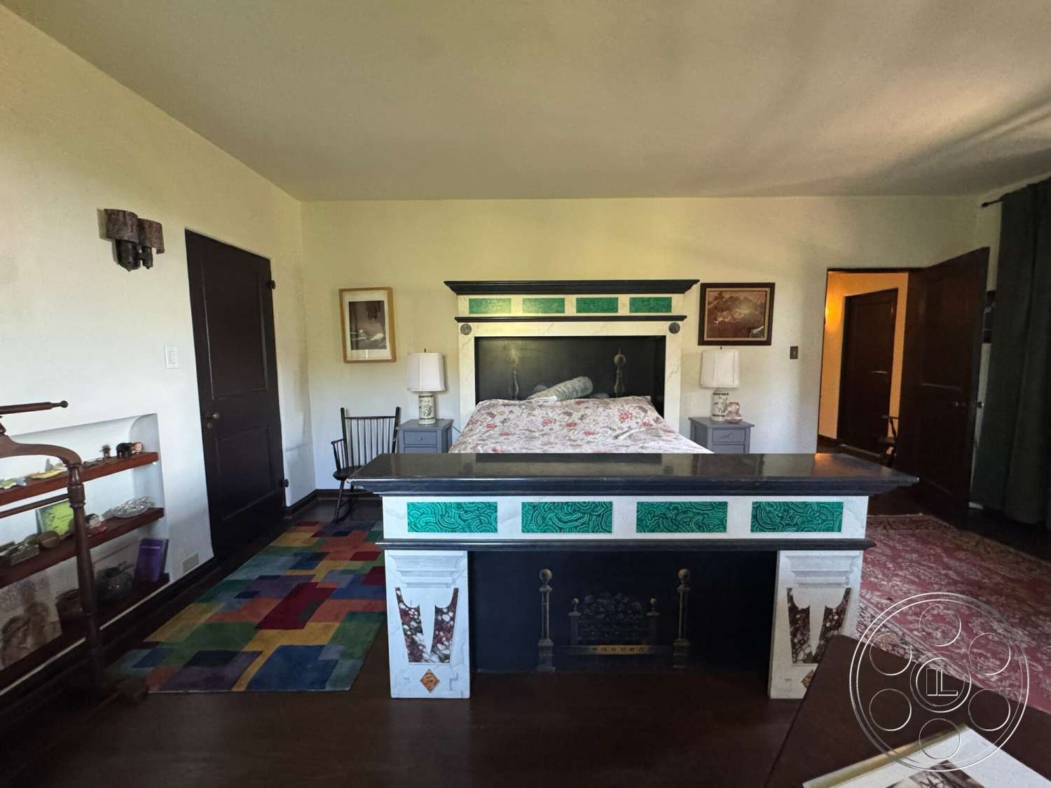 Farmstead Garden - artwork on walls, floor-to-ceiling windows, master bedroom, textured plaster walls, dark wood paneling, black wood furniture, colorful area rug, hardwood bedroom floors, white painted walls, built-in shelving unit, decorative fireplace surround, two doors, traditional bedside lamps, vintage chair