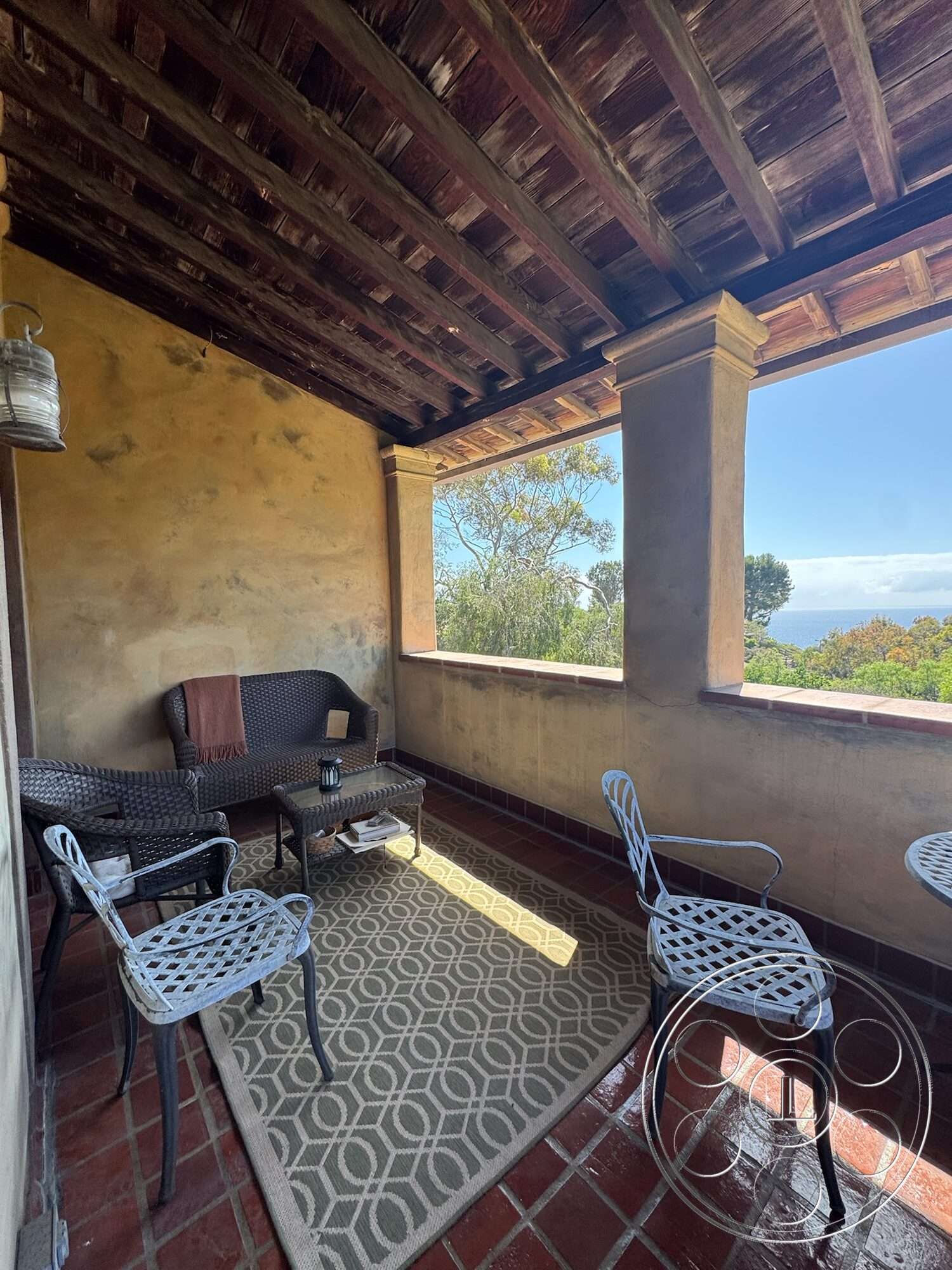 Farmstead Garden - exterior, textured area rug, terracotta tile flooring, wrought iron chairs, open-air seating area, natural landscape view, exposed wood ceiling beams, stucco wall treatment, woven wicker seating, open porch design