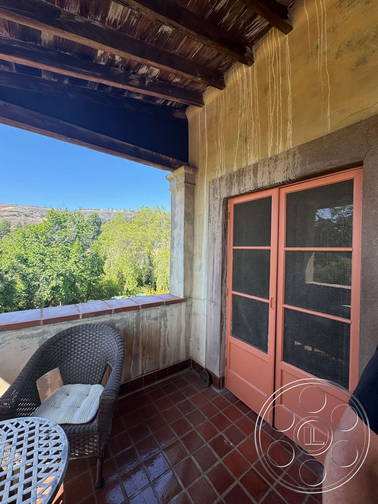 Farmstead Garden - exterior, balcony with railing, plastered walls, open-air patio, terracotta tile flooring, view of greenery, wood beam ceiling, pink wooden door, wicker patio chair