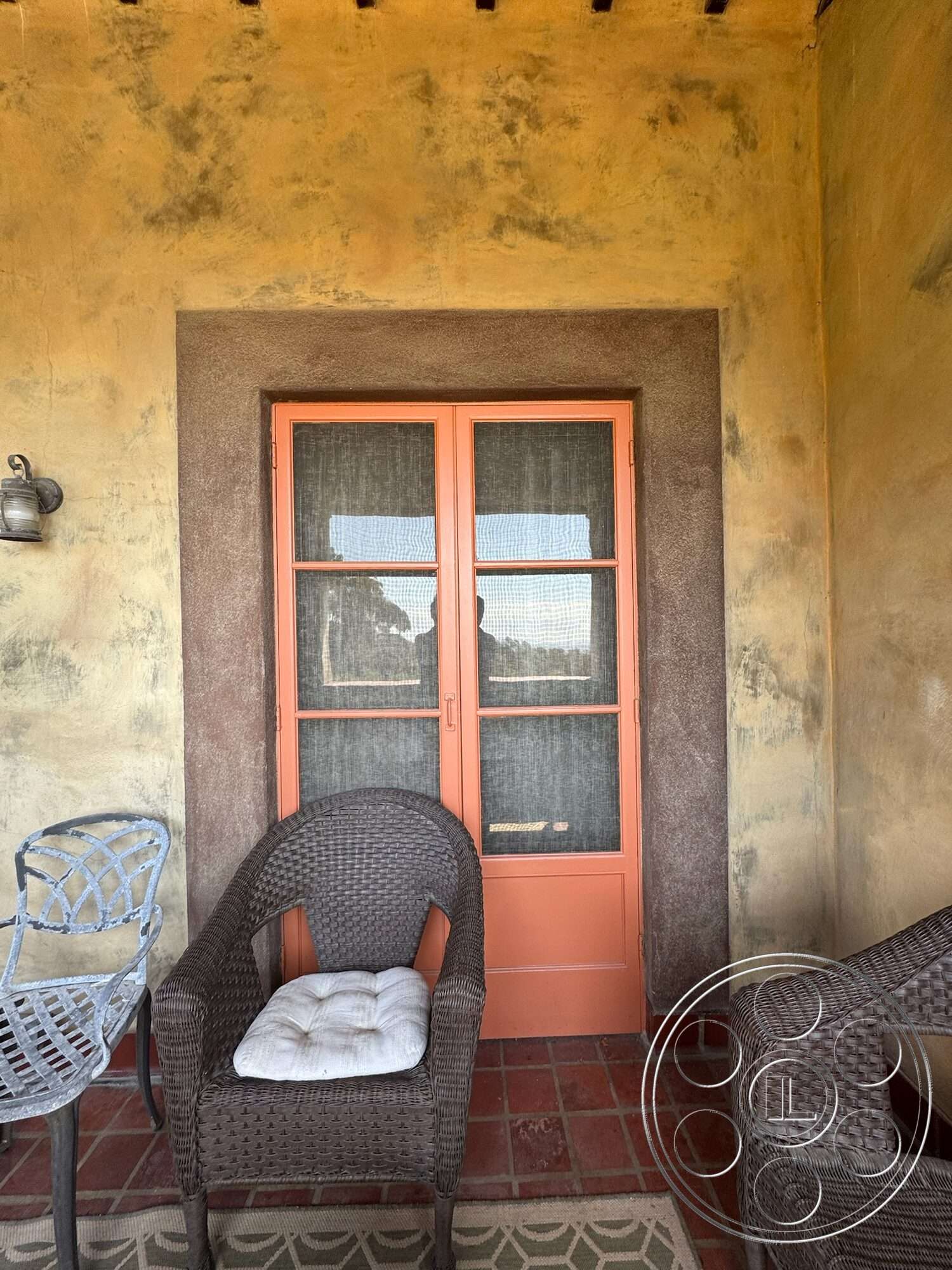 Farmstead Garden - exterior, outdoor seating area, stucco walls, decorative wall sconce, terracotta tile flooring, textured plaster wall, wood beam ceiling, brick patio border, woven patio chairs, orange double door, rustic patio, textile seat cushion