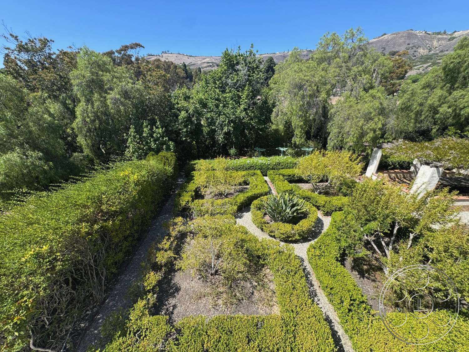 Farmstead Garden - exterior, landscaped garden, green shrubbery, flowering plants, mountain backdrop, clear blue sky, manicured hedges, gravel walking path, wooden garden benches, circular gravel garden beds, pathway edging stones, surrounding forest area