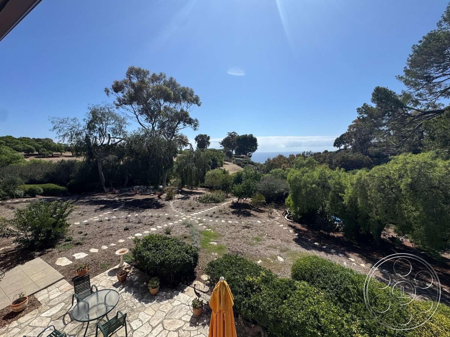 Farmstead Garden - outdoor living space, wooden patio furniture, drought-tolerant plants, sunny exterior, natural stone patio flooring, lush greenery landscaping, expansive ocean view, circular gravel garden path, umbrella shade in outdoor seating area, sloping terrain setting