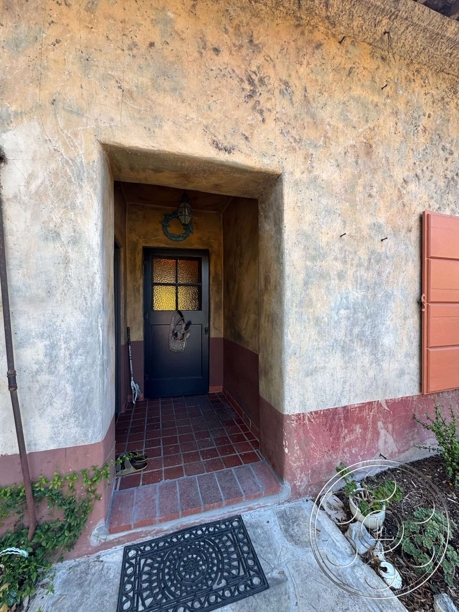 Farmstead Garden - exterior, arched doorway, welcome mat, landscaped garden area, textured plaster wall, stucco wall finish, red brick entryway flooring, green potted plants, earth-toned wall treatment, decorative wrought iron door, classic black door, terra cotta patio border, antique-style lantern light fixture