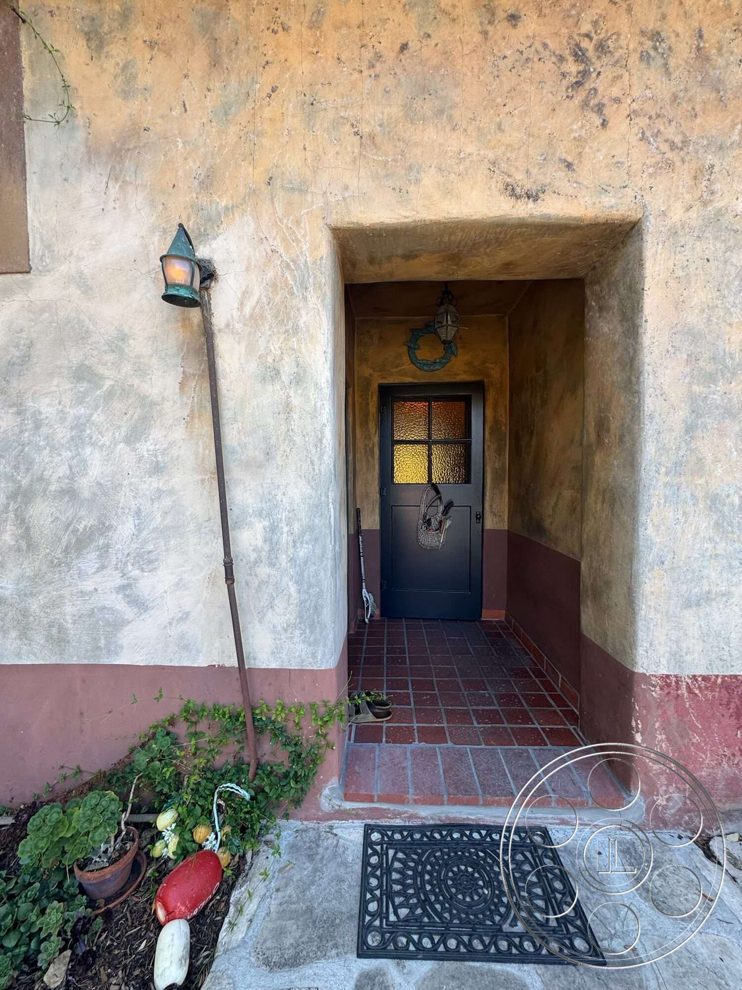 Farmstead Garden - exterior, glass panel door, textured plaster wall, stucco wall finish, dark wood entry door, landscaped entry garden, terra-cotta tile entryway floor, rustic wall sconce lighting, tile entry mat, ornamental metal door mat, exposed brick foundation, potted plants by entrance