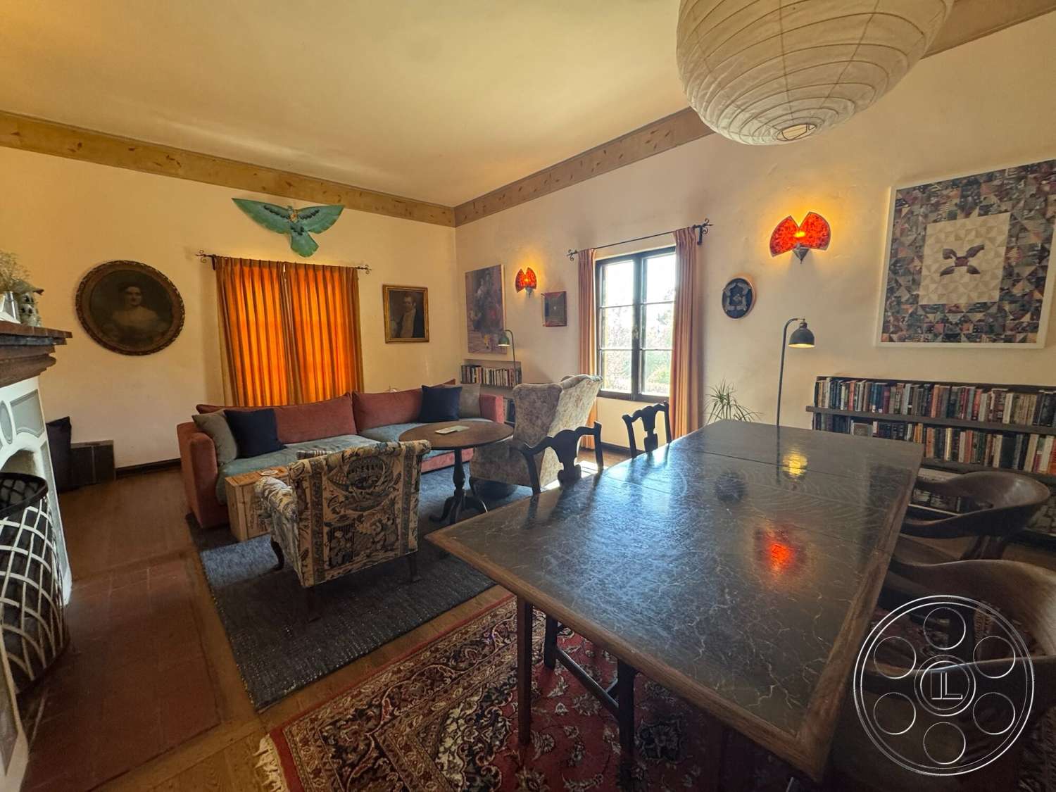 Farmstead Garden - tile flooring, green indoor plants, built-in bookshelf, woven area rug, textured plaster walls, decorative wall sconces, exposed brick fireplace surround, wood beam ceiling, orange sheer curtains, vintage artwork on walls, varnished wood dining table, fabric upholstered armchairs, ornate patterned upholstery