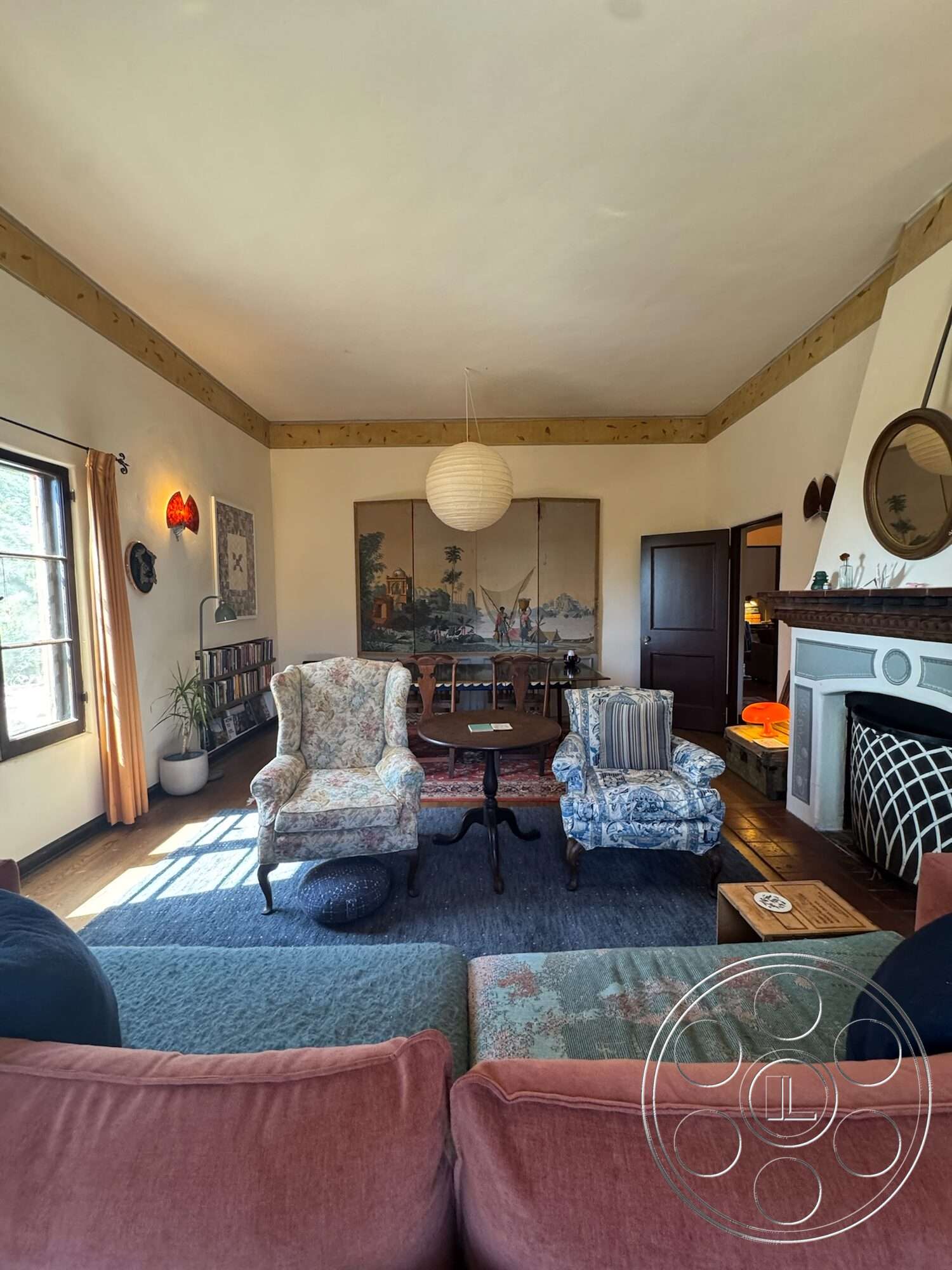 Farmstead Garden - hardwood flooring, wooden beam ceiling, floor-to-ceiling windows, soft area rug, textured plaster walls, round wooden coffee table, stone fireplace surround, exposed wood ceiling, vintage wall sconces, blue floral upholstered armchairs, oriental decorative panels, earth-toned curtains, circular paper lantern light fixture, three-seater sofa with fabric upholstery