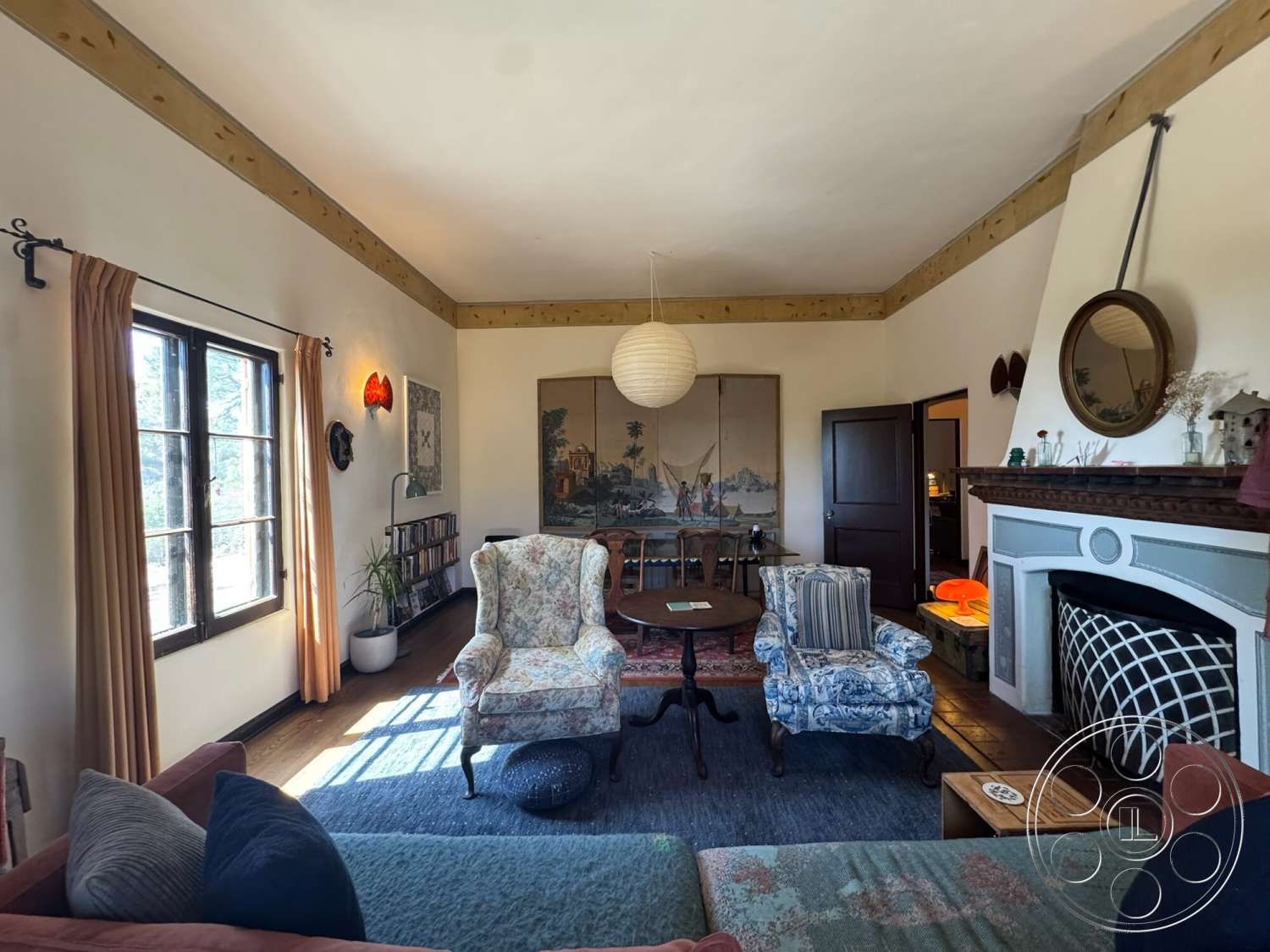 Farmstead Garden - floor-to-ceiling windows, decorative wall art, oriental rug, dark wood door, beige curtains, exposed wood ceiling beams, hardwood living room floors, textured plaster wall, potted indoor plants, floral patterned armchair, traditional-style chandelier, light-colored painted walls, striped armchair, wooden round coffee table, fireplace with ornamental mantle, vintage wall sconce, traditional-style decor