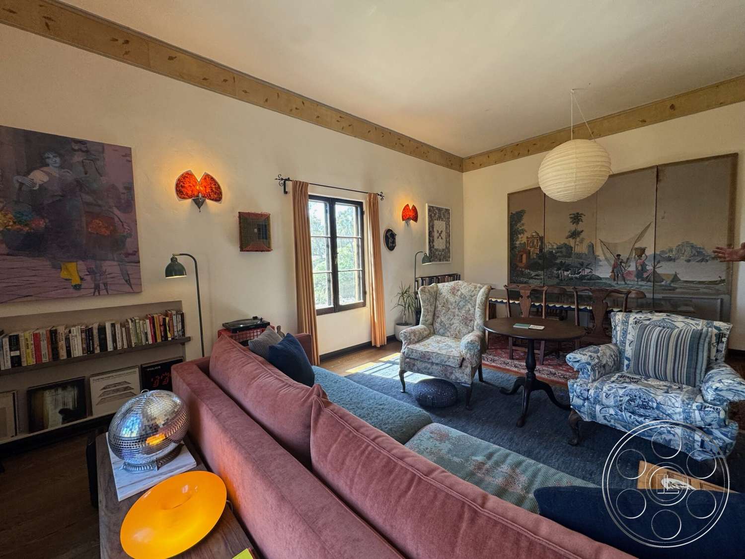Farmstead Garden - hardwood flooring, indoor plant, area rug, decorative wall art, textured plaster walls, round wooden coffee table, wood beam ceiling, large windows with curtains, pink upholstered sofa, striped armchair, floral armchair, wall-mounted lamps, overhead paper lantern light, book shelving unit, multi-colored throw pillows