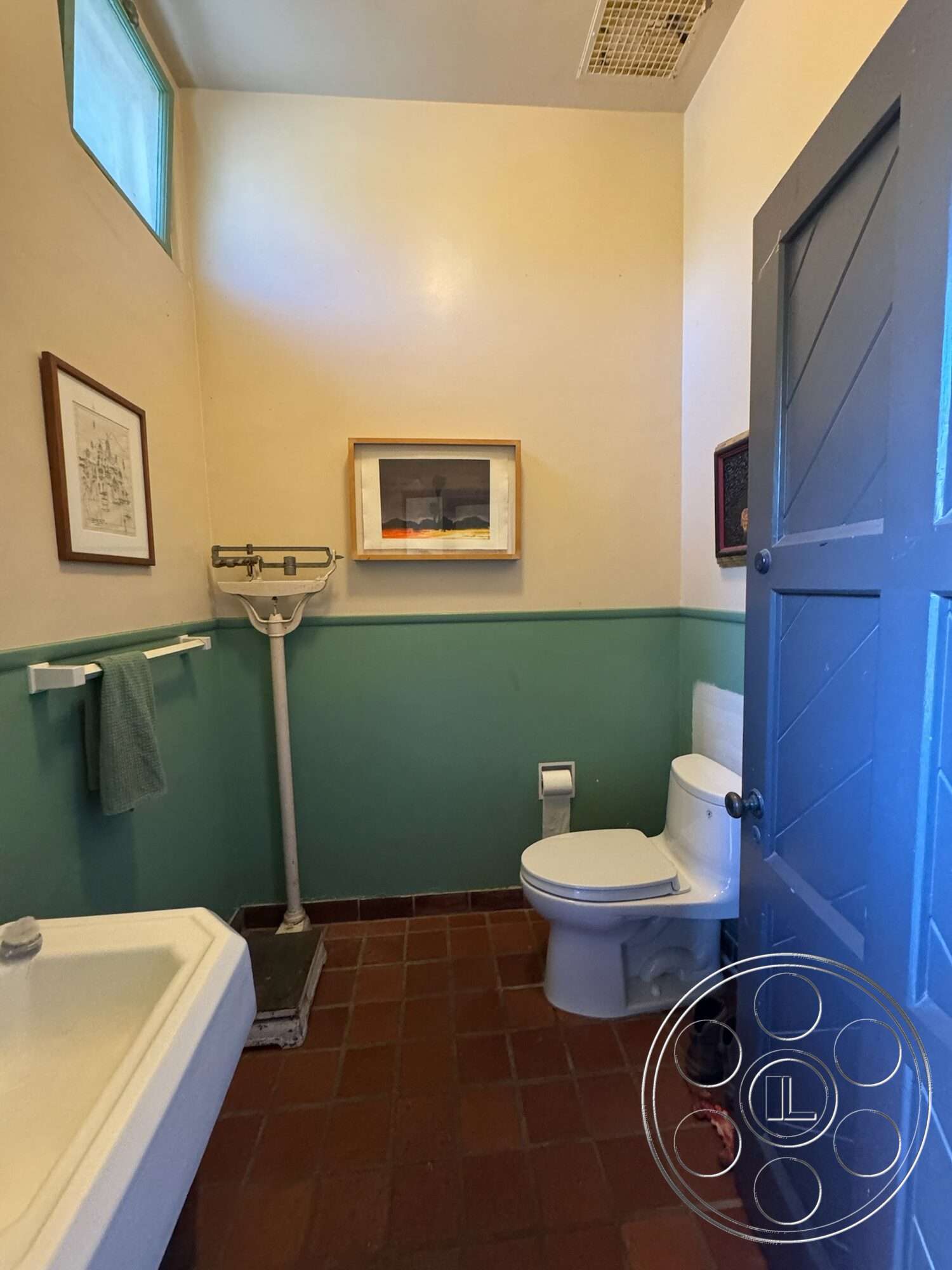 Farmstead Garden - framed wall art, textured plaster walls, green accent wall, white porcelain toilet, terracotta tile flooring, blue wooden door, cream painted walls, white bathroom sink, wooden framed artwork, metal towel rack, classic door handle