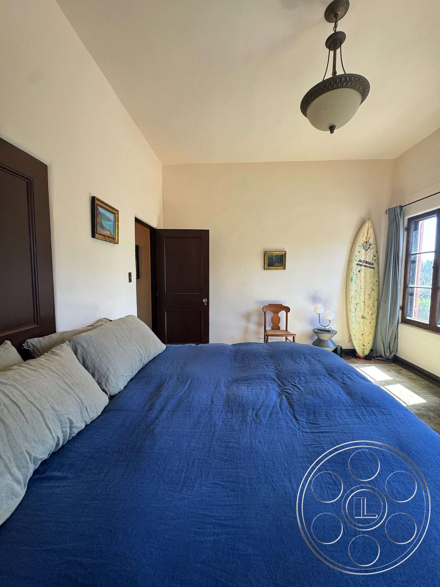 Farmstead Garden - neutral color palette, natural light, table lamp, cozy atmosphere, floor-to-ceiling windows, vaulted ceiling, textured plaster walls, dark wood paneling, wooden chair, green velvet bedding, surfboard wall art