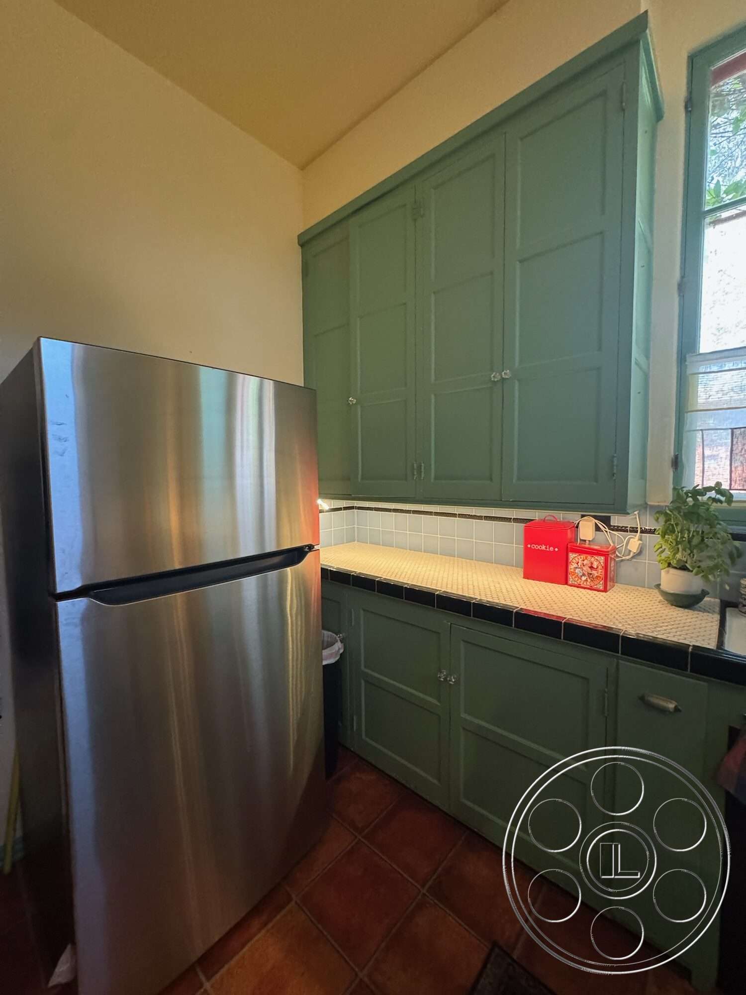 Farmstead Garden - stainless steel refrigerator, double-hung windows, interior kitchen, green painted kitchen cabinets, white tile countertops, black tiled backsplash, terra-cotta kitchen flooring