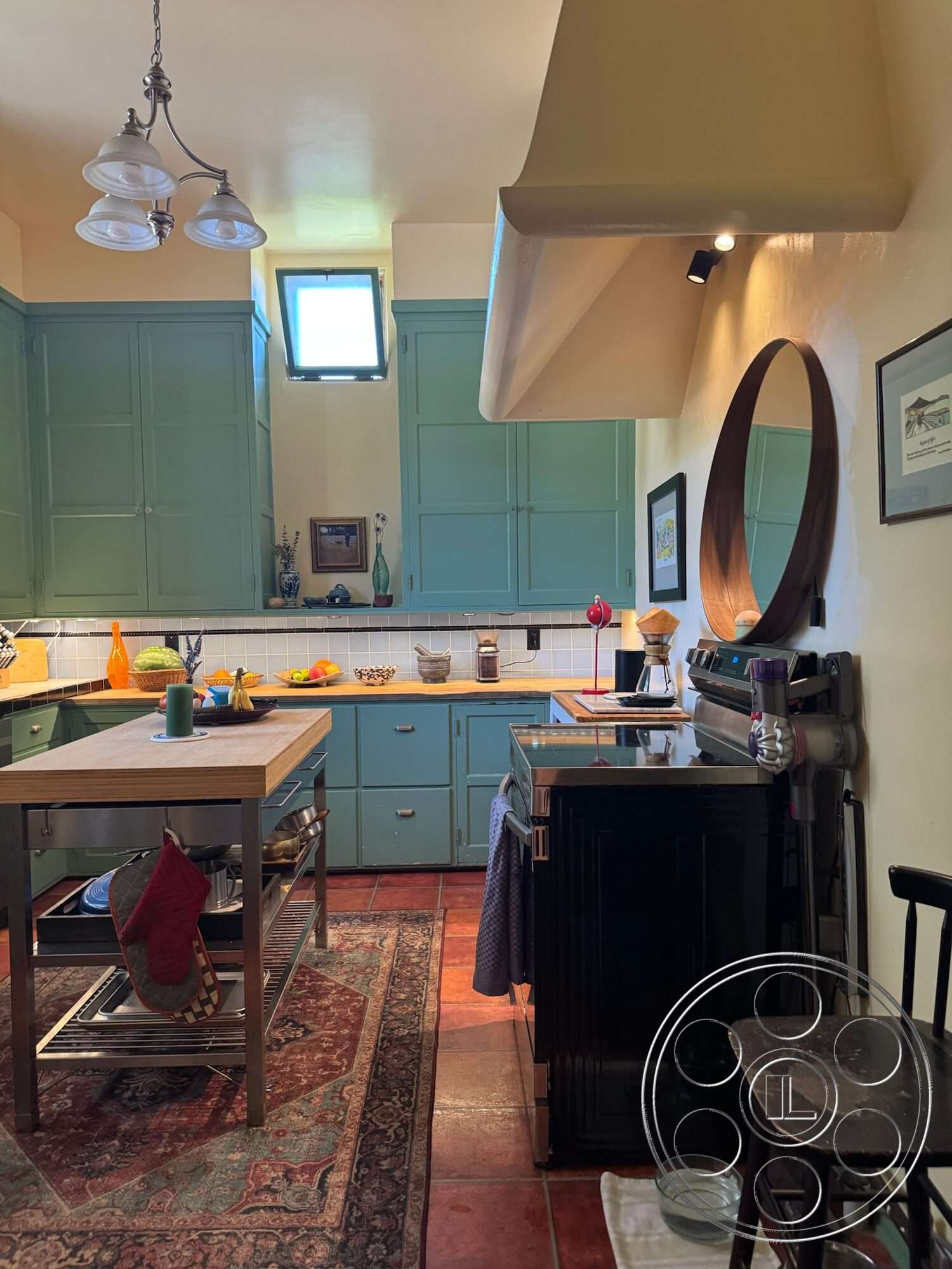 Farmstead Garden - decorative fruit bowl, stainless steel kitchen appliances, exposed ductwork ceiling, pendant light fixtures, rustic area rug, white tiled kitchen backsplash, green painted kitchen cabinets, terra cotta tile kitchen floors, wood countertop kitchen island, black enamel kitchen cabinetry, round mirror wall decoration