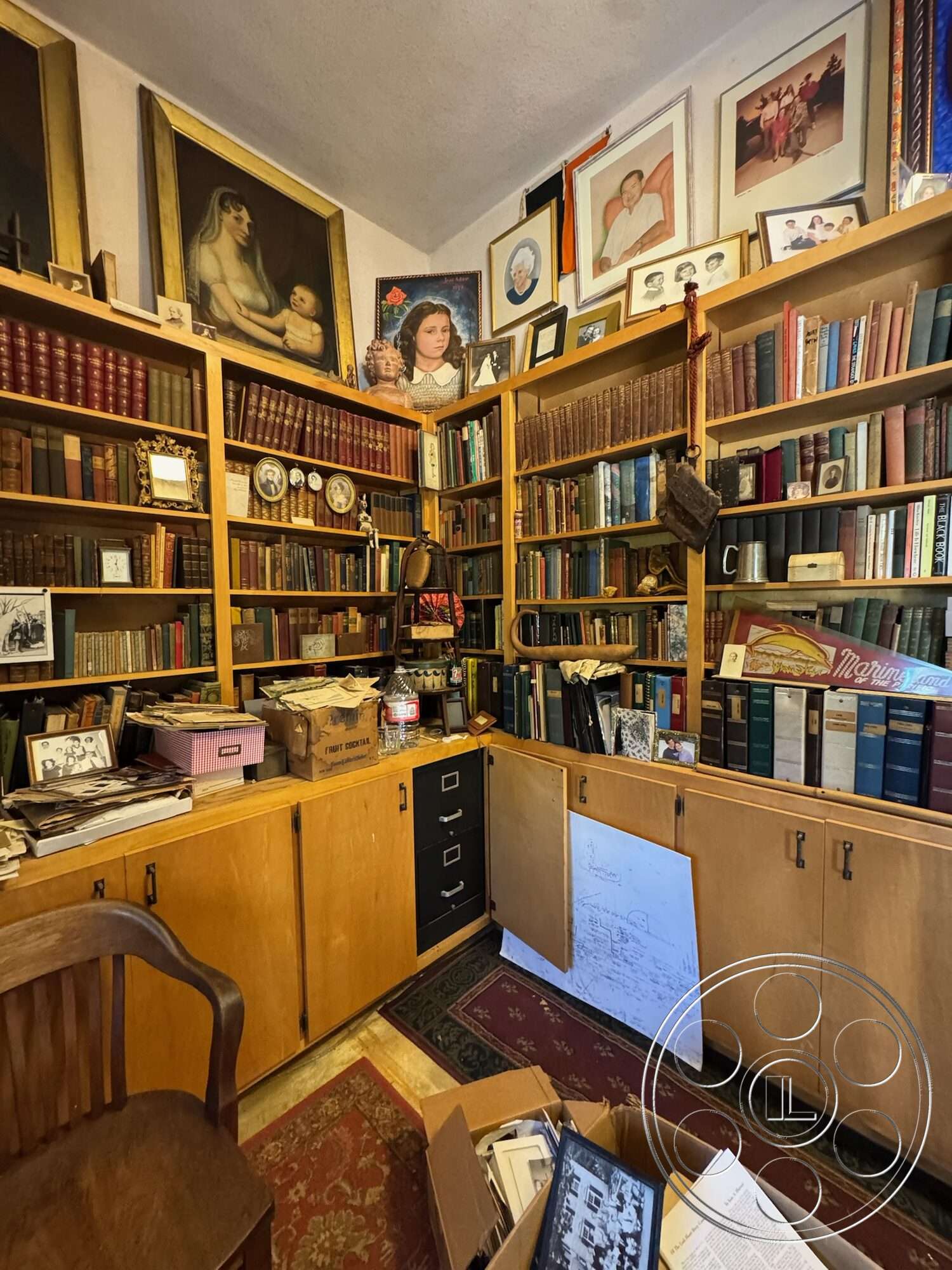 Farmstead Garden - white plaster walls, framed artwork on walls, vintage wooden chair, wooden bookshelves filled with books, light wood cabinets with metal handles, oriental rug flooring, box of documents on the floor, cluttered workspace with various papers and boxes