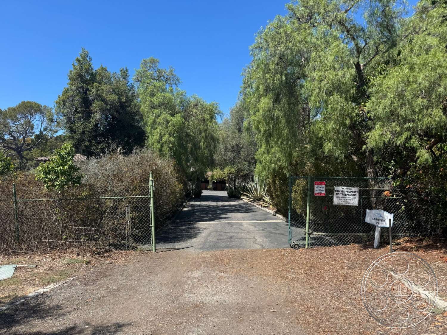 Farmstead Garden - paved driveway, lush greenery, mature trees, gated entrance, clear blue sky, private property, gravel landscape, chain link fence, sunny outdoor setting