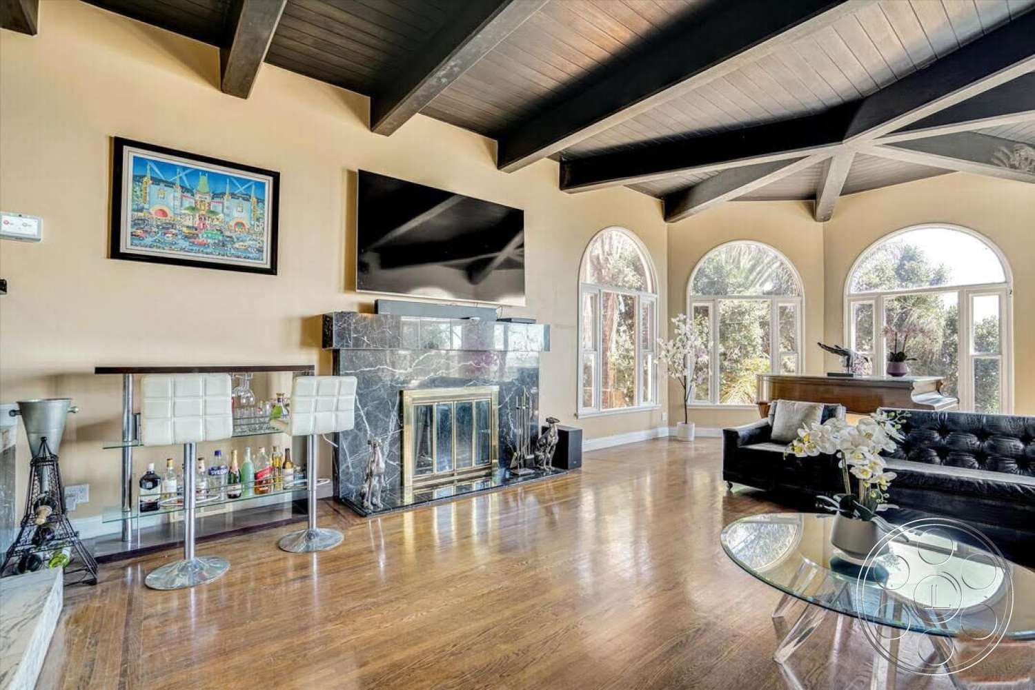 Spanish 170 - glass coffee table, decorative wall art, natural light from large windows, modern television, arched windows, black leather sofa, exposed wood ceiling beams, hardwood living room floors, neutral wall paint, marble fireplace surround, chrome barstools, glass bar cart