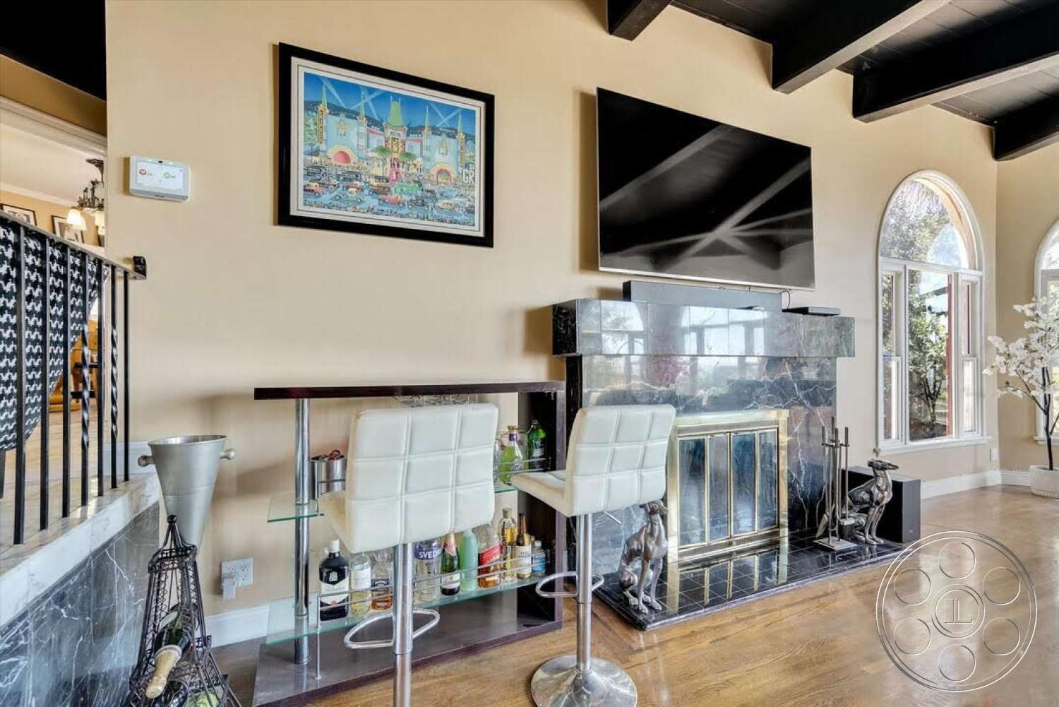 Spanish 170 - framed wall art, arched windows, large flat-screen television, dark wood ceiling beams, hardwood living room floors, tan painted walls, decorative plant accents, black marble fireplace surround, white leather bar stools, glass bar shelf