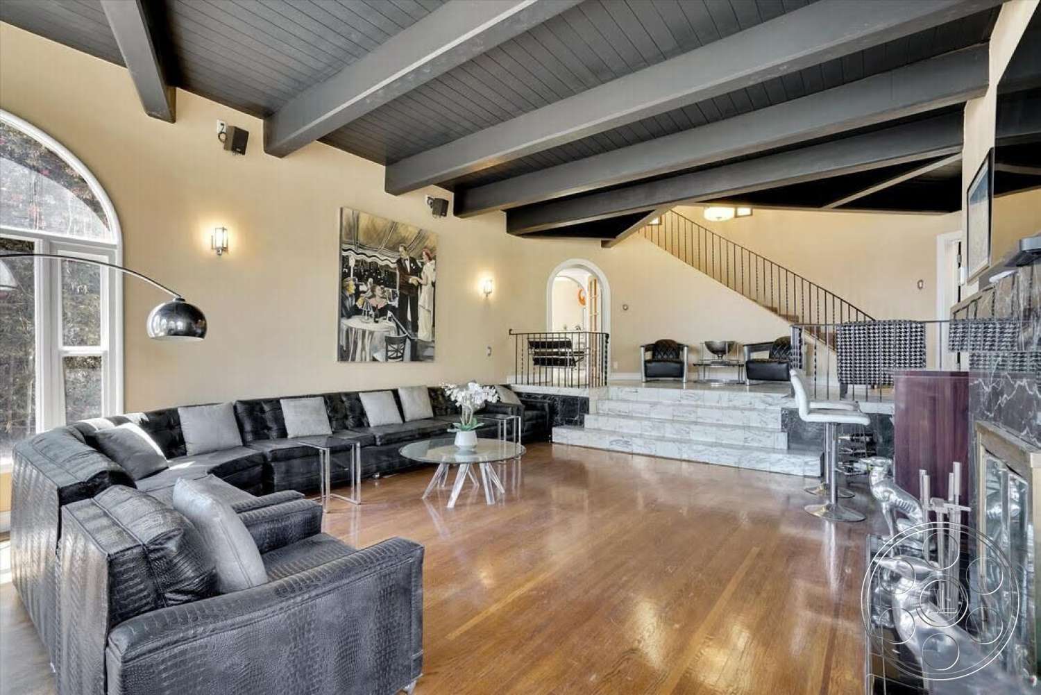Spanish 170 - metal railing, artwork on wall, vaulted ceiling, large arched windows, curved staircase, round glass coffee table, wall-mounted light fixtures, exposed wood ceiling beams, hardwood living room floors, neutral painted walls, stone fireplace wall, black leather sectional sofa, marble steps