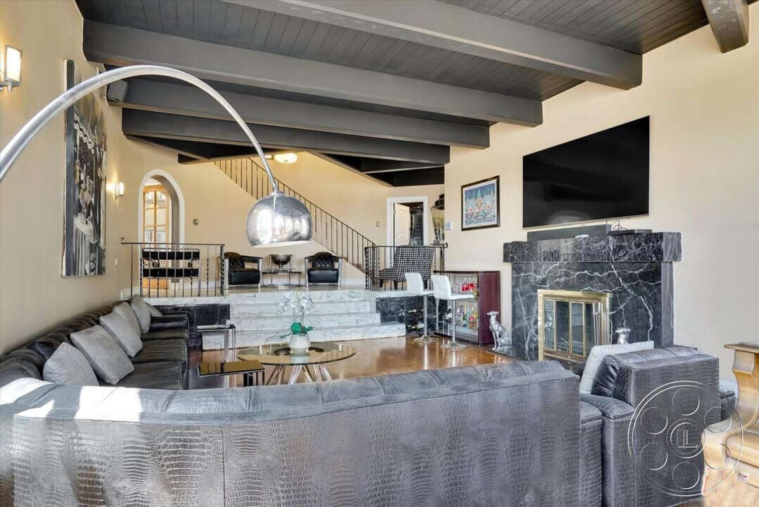 Spanish 170 - open floor plan, glass coffee table, dark gray sectional sofa, arched doorways, flat-screen television, stylish accent chairs, exposed wood ceiling beams, hardwood living room floors, neutral wall paint, marble fireplace surround, metal stair railing, contemporary wall art