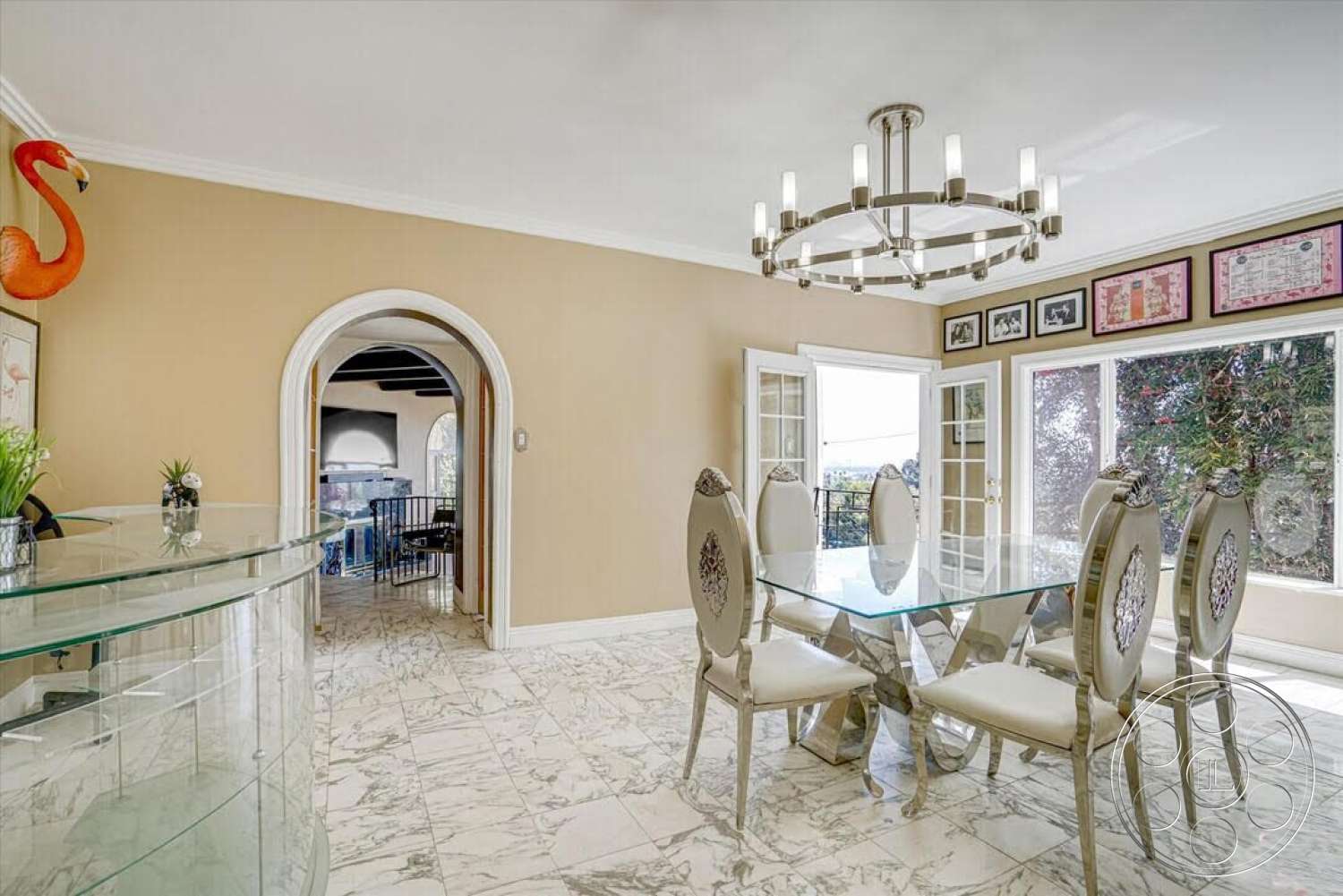 Spanish 170 - plant decor, open floor plan, glass dining table, decorative wall art, arched doorways, exposed wood ceiling beams, neutral painted walls, crown molding details, chandelier lighting fixture, large picture windows, marble dining room flooring, metallic dining chairs