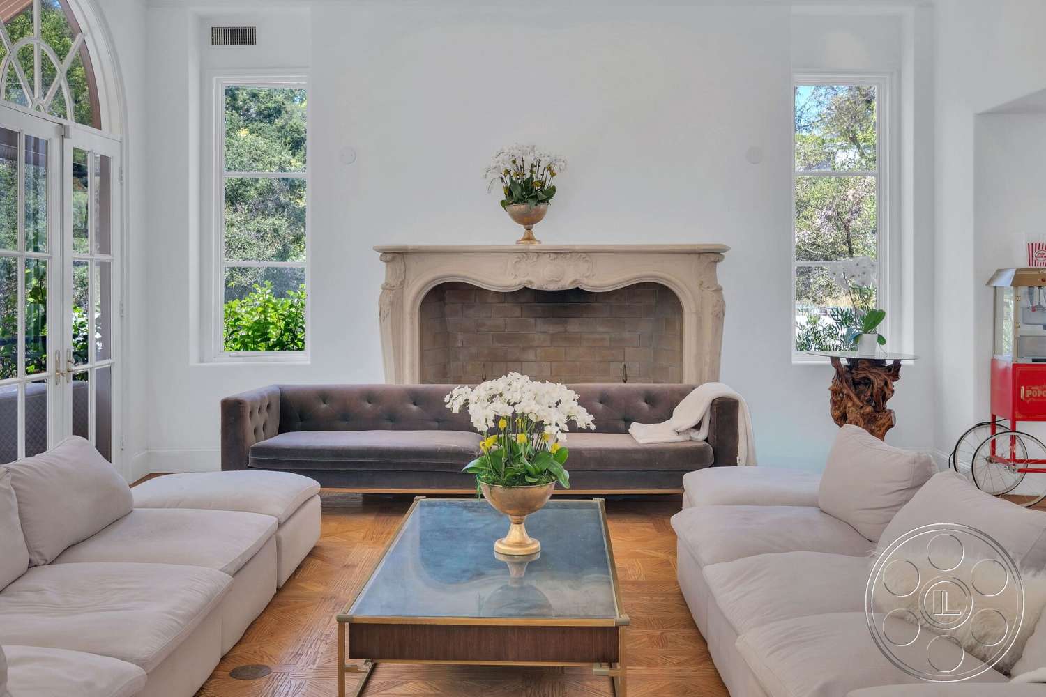 French 29 - natural light, glass coffee table, floor-to-ceiling windows, lush greenery view, exposed brick fireplace wall, white painted walls, arched window frames, decorative ceiling molding, hardwood herringbone living room flooring, modern tufted sofa, stylish decorative accents