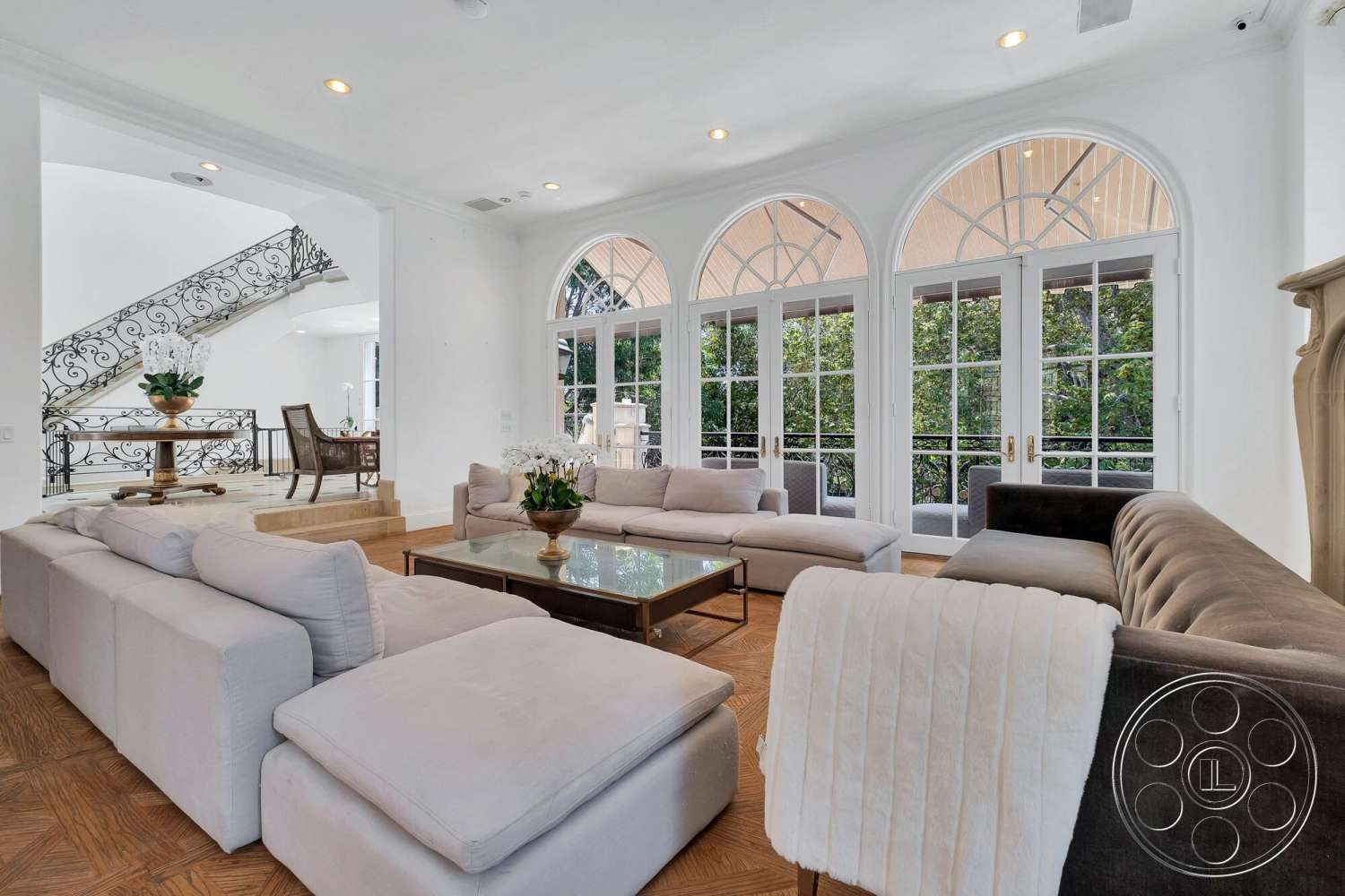French 29 - neutral color palette, glass coffee table, plush sectional sofa, flat ceiling with recessed lighting, open floor plan design, herringbone hardwood living room floors, decorative fireplace surround, stainless steel decorative railing, arched windows with grid pattern, round ornamental planter with flowers, expansive natural light from windows