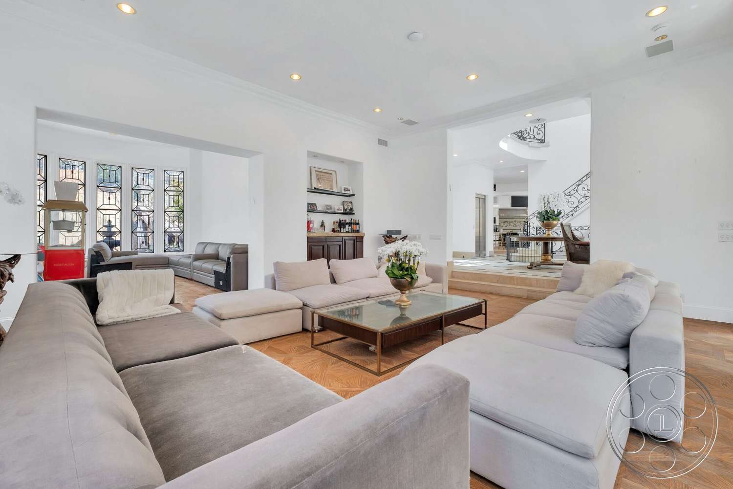French 29 - glass coffee table, white painted walls, curved staircase with wrought iron railing, decorative plant arrangement, hardwood herringbone living room floors, sofa with light grey upholstery, arched window with stained glass, shelves with built-in cabinetry