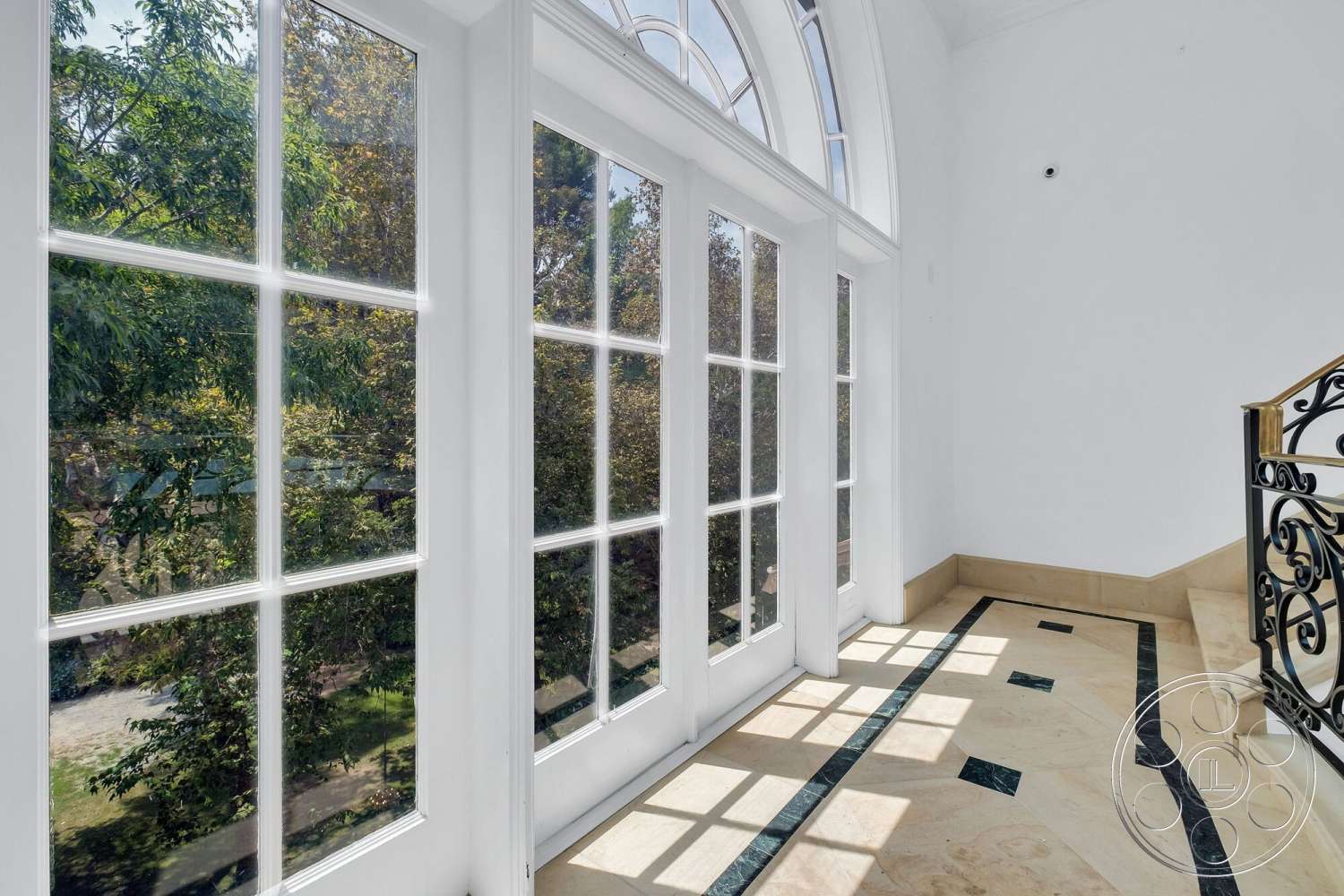 French 29 - high ceiling, floor-to-ceiling windows, curved staircase, white painted walls, natural light exposure, elegant entryway design, marble flooring with contrasting stone inlays, ornate metal stair railing, lush greenery view through windows
