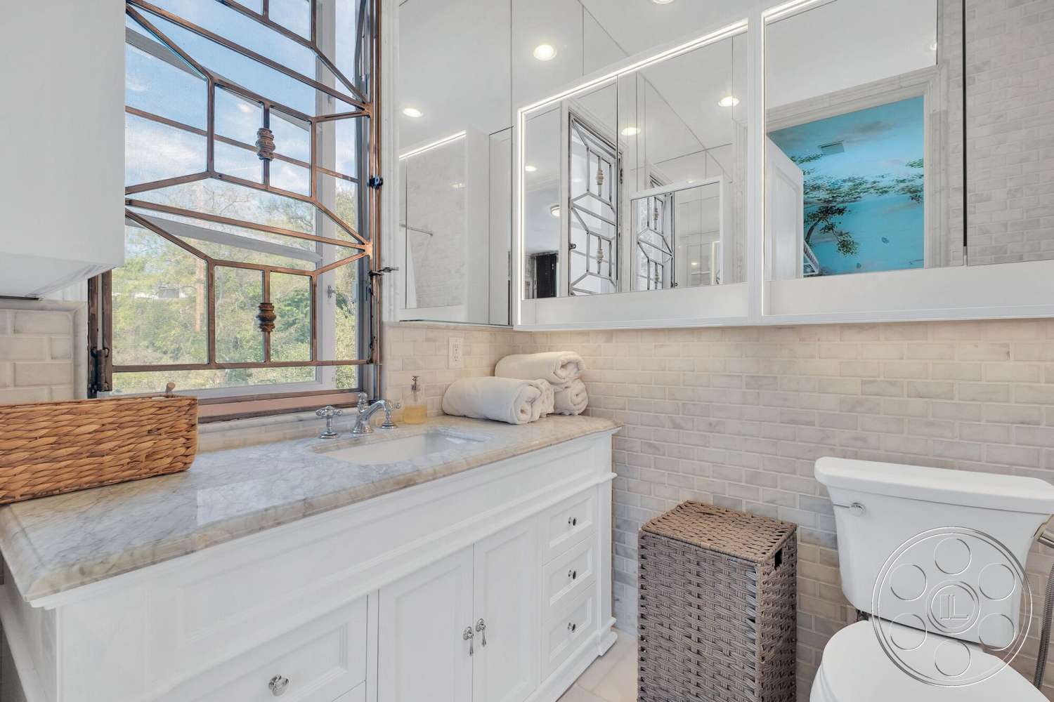 French 29 - subway tile backsplash, white cabinetry, marble countertop, woven storage basket, framed mirror wall, textured beige tile walls, glass window with metal frame, decorative wicker basket, ceramic toilet