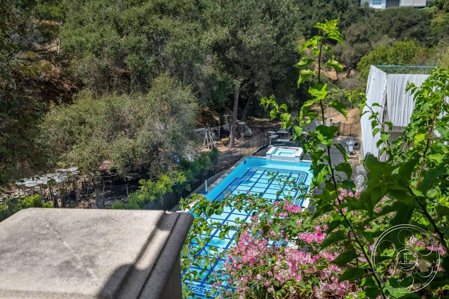 French 29 - exterior, surrounding greenery, sloped terrain, mountain backdrop, concrete pool deck, rectangular pool, wooden patio furniture, stone landscaping, fenced backyard, decorative flowering bushes, pool safety cover