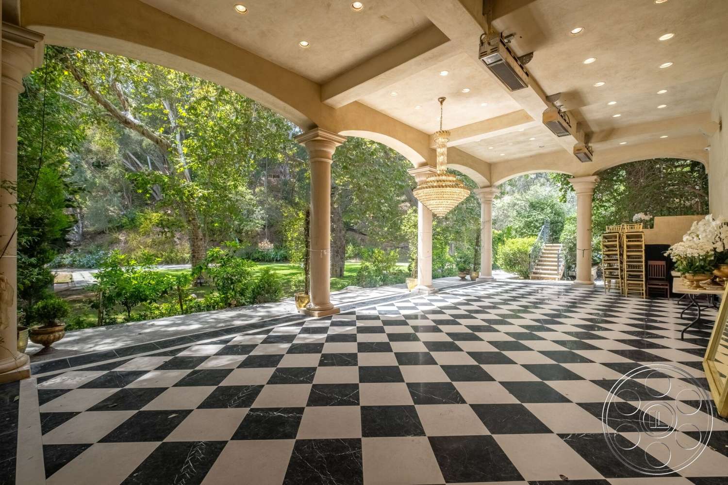 French 29 - exterior, decorative planters, textured plaster ceiling, modern outdoor space, chandelier lighting fixture, arched entryway with columns, polished marble flooring in black and white checkerboard pattern, lush greenery in background, paved stone pathway beyond entry, surrounding garden landscaping