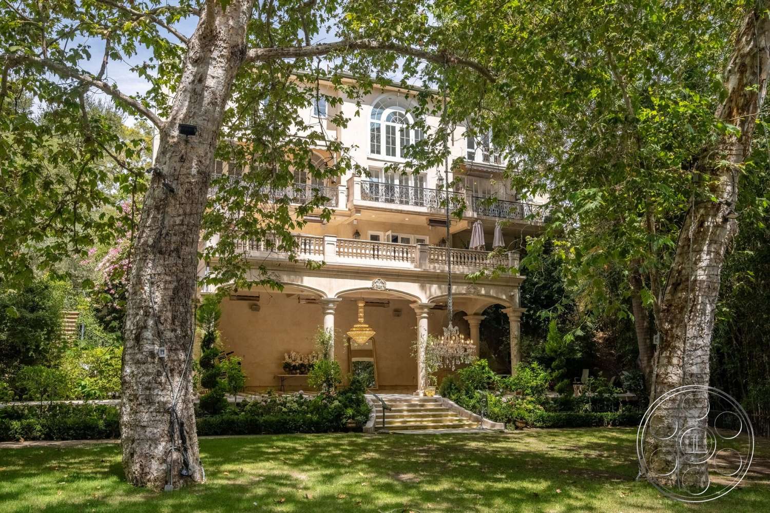 French 29 - exterior, stucco facade, arched entryway, tile roof, stone steps, balcony with wrought iron railings, decorative stone planters, landscaped garden area, mature trees surrounding property, large multi-paned windows, detailed column support, chandelier hanging from covered porch, flat yard area