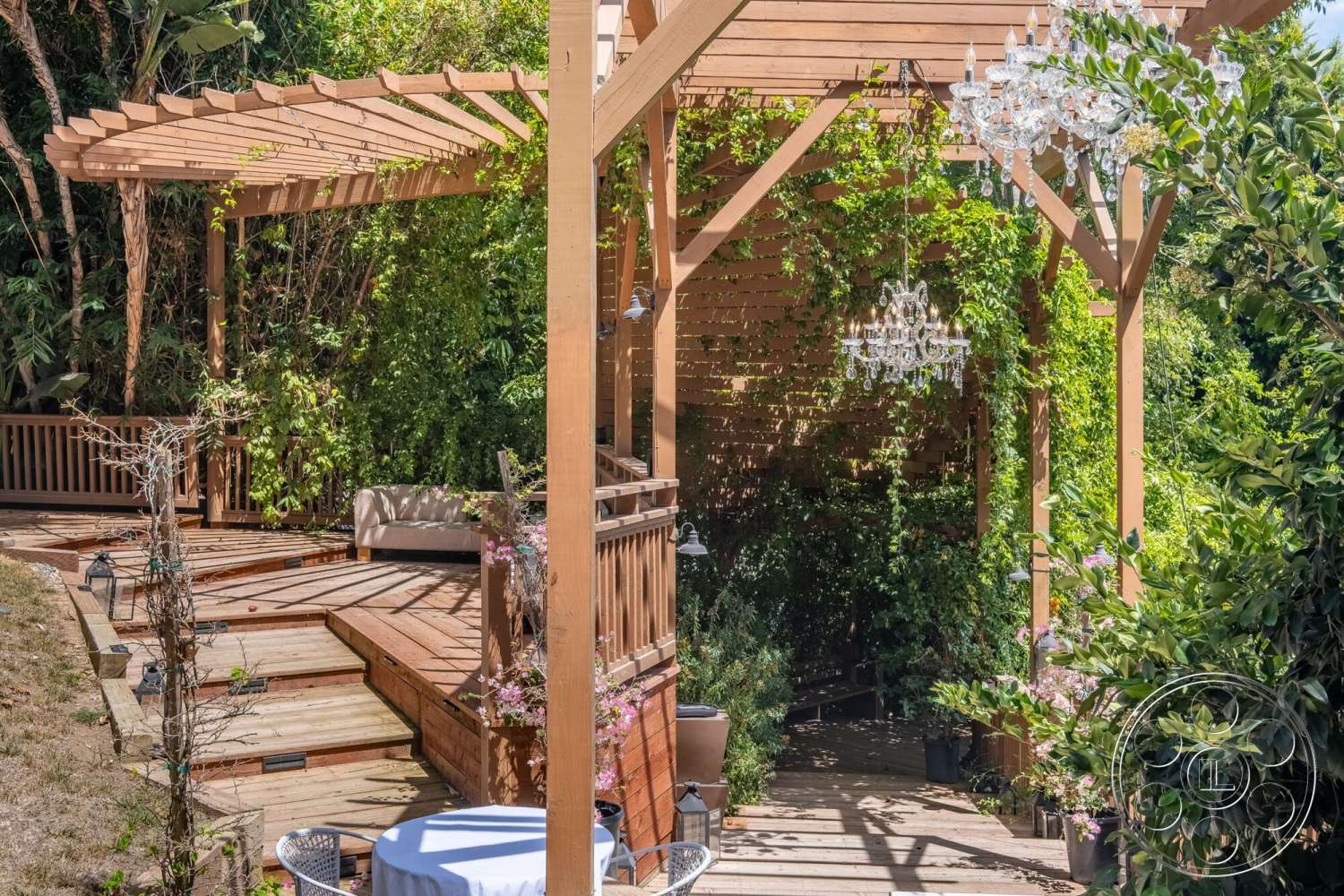 French 29 - exterior, outdoor seating area, planted greenery, decorative potted plants, lush green landscaping, wooden deck flooring, wood pergola structure, wooden railing details, hanging crystal chandeliers, staircase with wooden treads