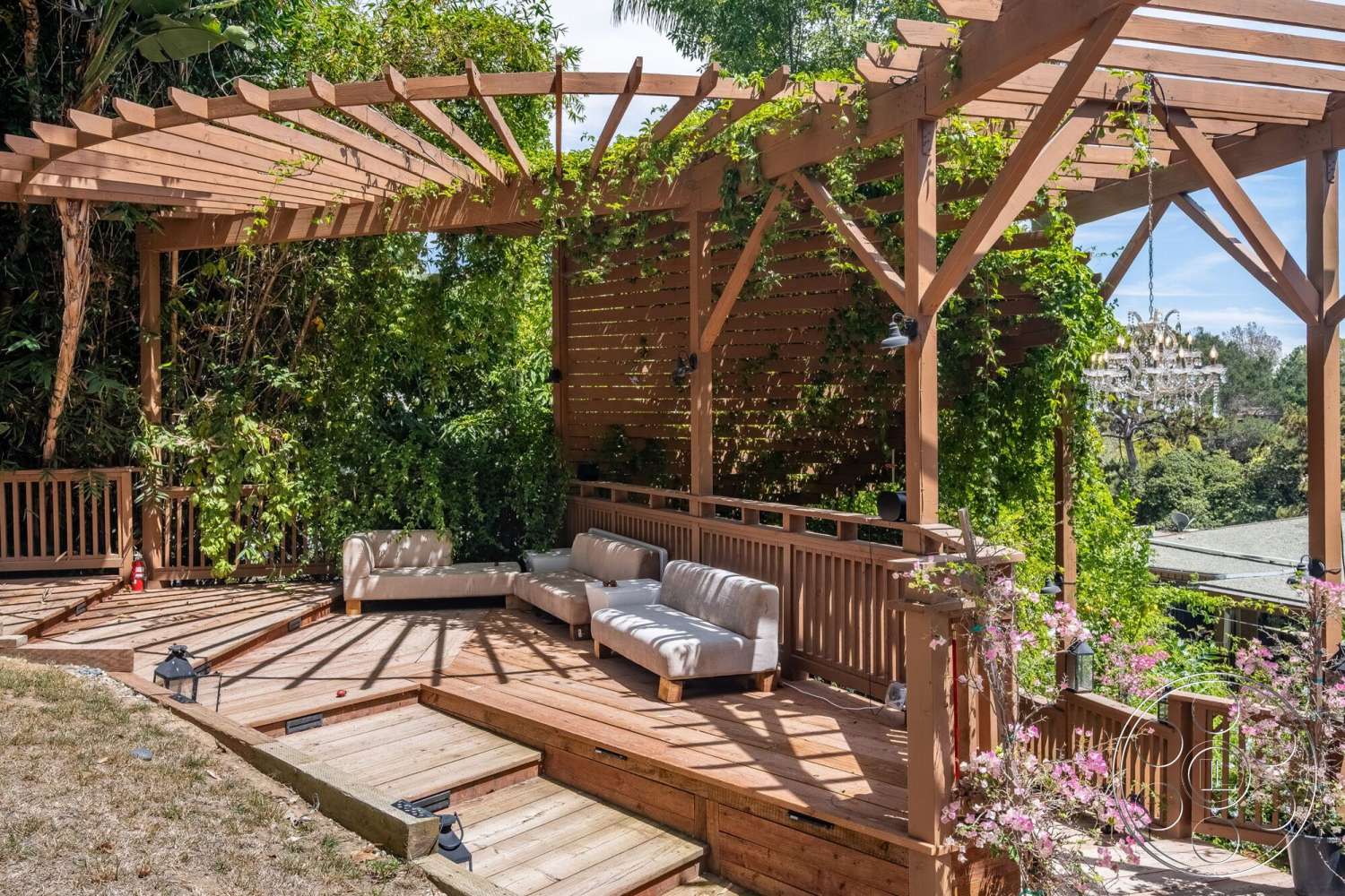 French 29 - exterior, wooden decking, stone pathway, wooden pergola, garden patio, wooden railing, lush green landscaping, fabric outdoor cushions, modular outdoor seating, cedar wood beams, trellis with climbing vines, chandelier hanging from pergola