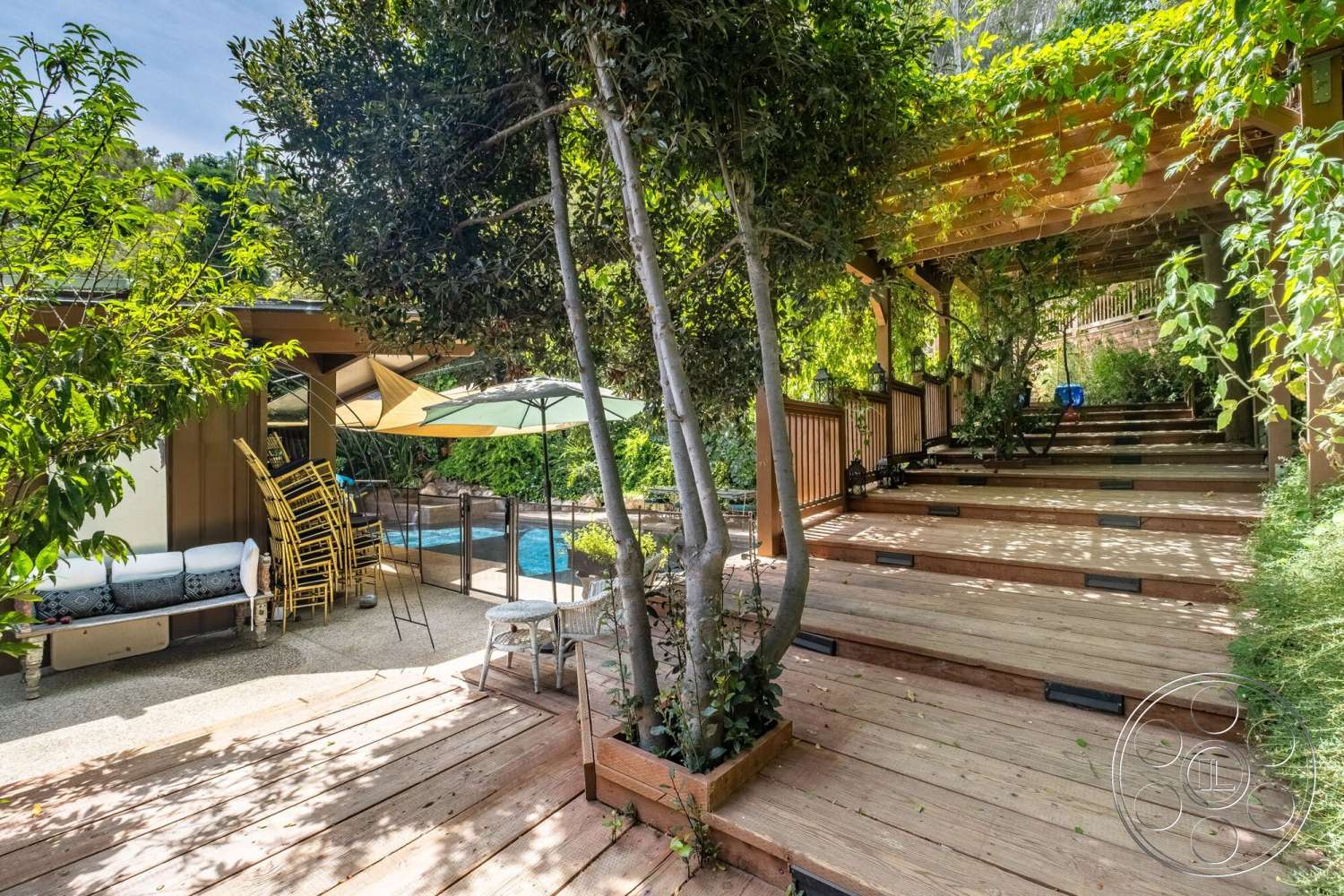 French 29 - exterior, surrounding trees, umbrellas, wooden pergola, in-ground swimming pool, landscaped garden area, wooden deck steps, concrete patio floor, planter boxes with greenery, stacked wooden chairs