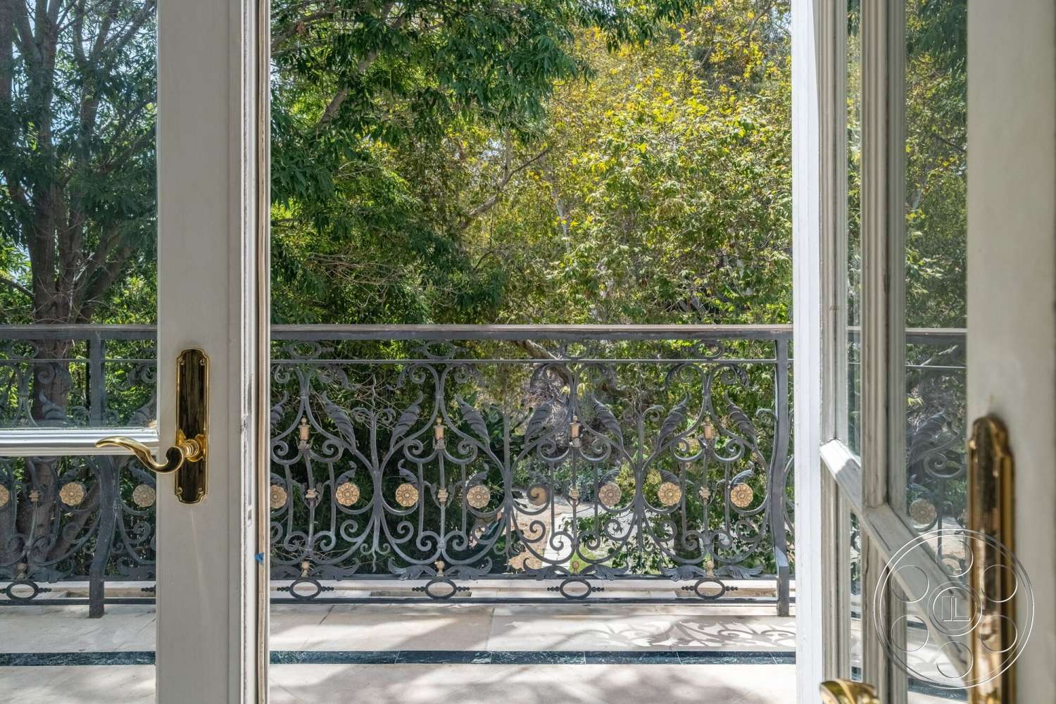 French 29 - natural light filtering through foliage, exterior balcony railing with ornate wrought iron design, framed glass door with polished brass handle, marble balcony flooring, lush greenery with trees in the background, outdoor view from a second-story balcony, elevated balcony setting