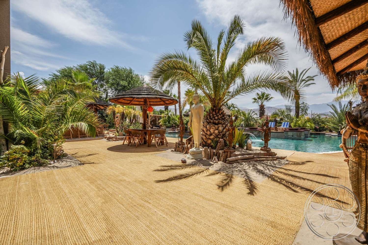 Rancho Mirage 4 - exterior, thatched roof gazebo, rectangular pool, stone pool deck, wooden outdoor dining table, tropical palm landscaping, mountain view backdrop, warm natural lighting, decorative garden sculptures, woven straw patio flooring, sand feature around pool
