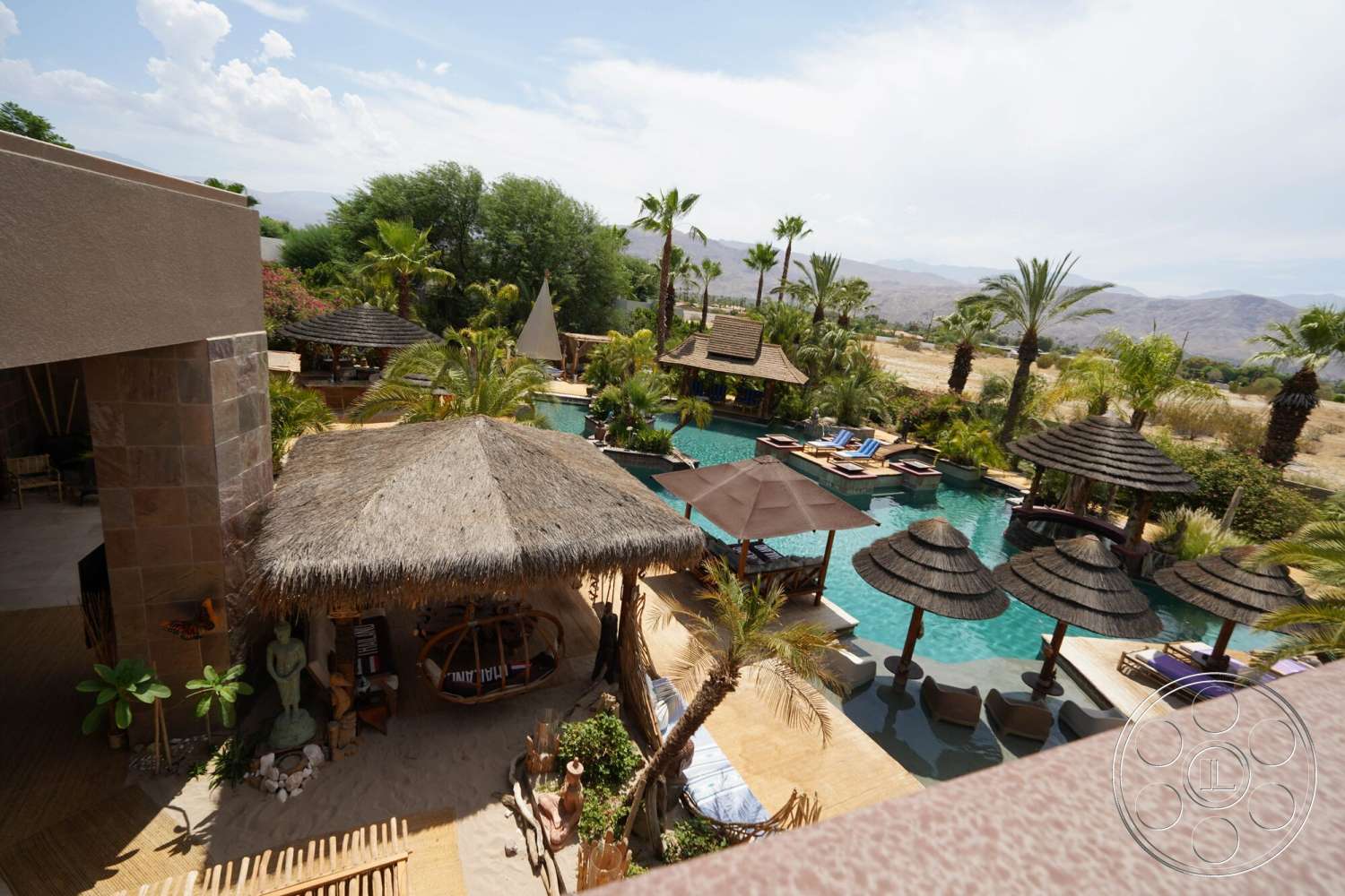 Rancho Mirage 4 - exterior, outdoor seating area, infinity pool, natural stone wall, concrete pool deck, sunshade umbrellas, wooden lounge chairs, palm tree landscaping, tile pool flooring, desert landscape setting, resort-style outdoor area, decorative garden sculptures, sandstone patio flooring, thatched roof cabanas