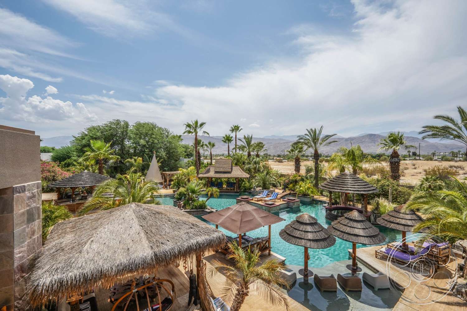 Rancho Mirage 4 - exterior, outdoor seating area, rectangular swimming pool, mountain view, relaxation zone, concrete pool coping, palm tree landscaping, wooden patio deck, desert backdrop, sunny sky, thatched roof cabanas, tropical garden area, beach lounge chairs, natural stone material