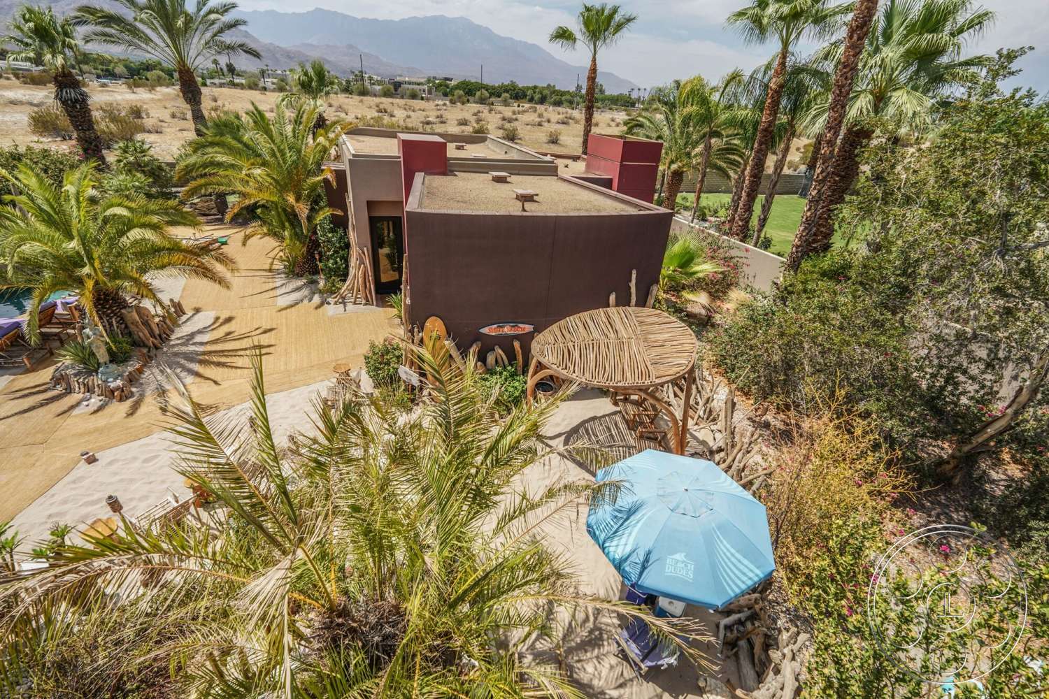 Rancho Mirage 4 - exterior, flat roof, concrete walkway, desert landscape, stucco walls, tropical palm trees, planting beds, wooden lounge chairs, rectangular patio, scenic mountain view, blue patio umbrella, wooden outdoor table