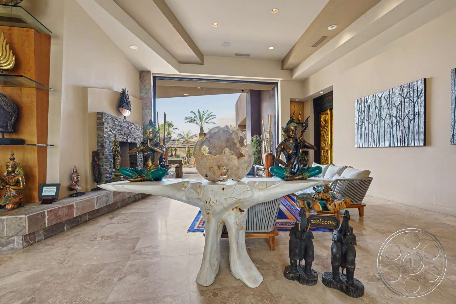 Rancho Mirage 4 - neutral wall colors, open concept living space, large sliding glass doors, contemporary coffee table, high ceilings with recessed lighting, modern wall art, elegant decorative accents, travertine floor tiles, natural stone fireplace wall, decorative stone shelving, ornamental sculptures, tropical outdoor view