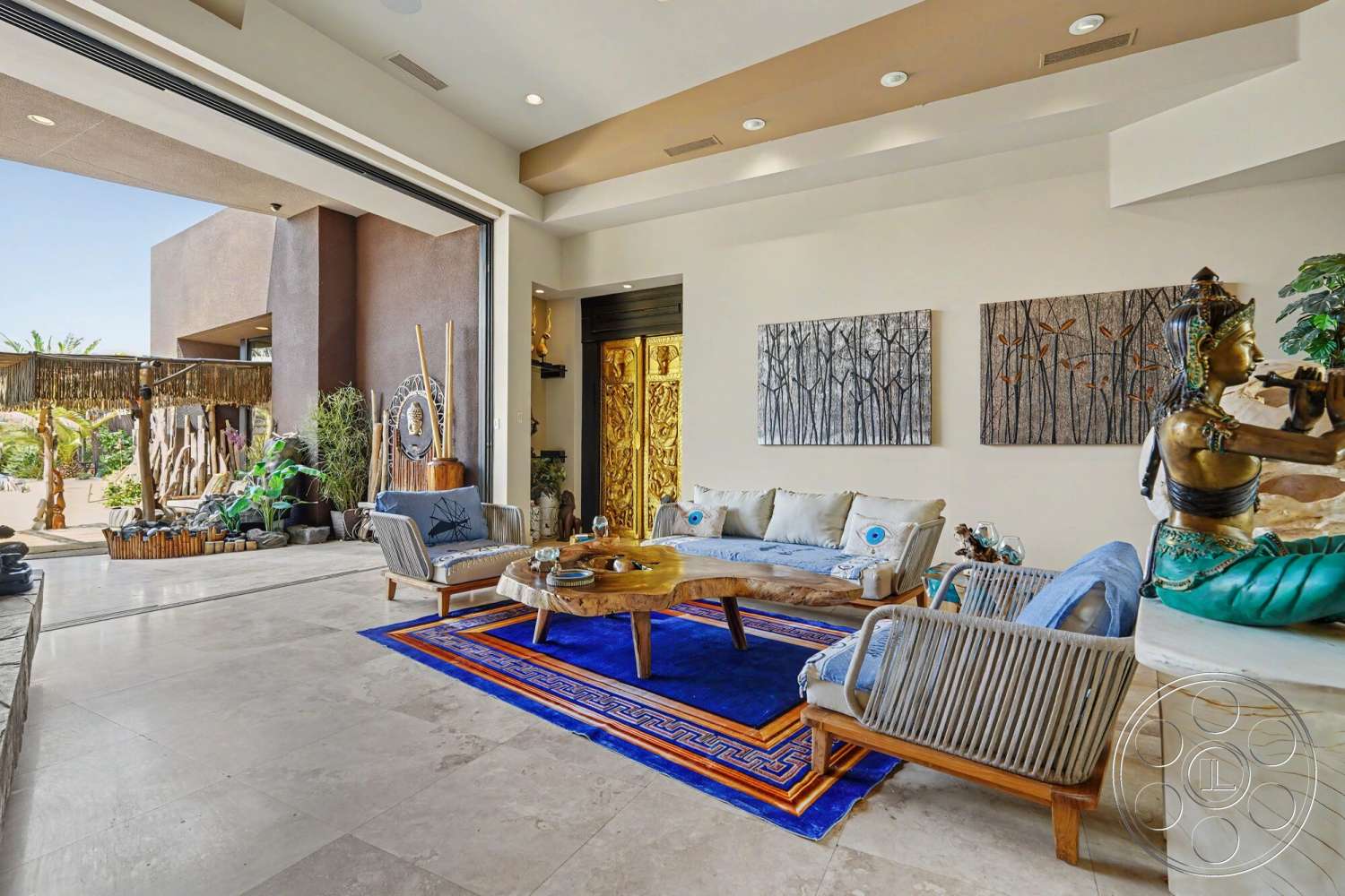 Rancho Mirage 4 - high ceiling with recessed lighting, neutral painted walls, potted indoor plants, tile living room flooring, open floor plan with natural light, natural stone coffee table, folding glass wall opening, wood accent wall decor, bohemian style seating, handcrafted wooden side tables, textile area rug with geometric patterns, large wall art with forest theme, golden ornamental door, artistic figurine display
