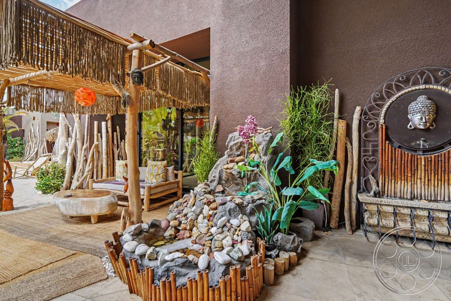 Rancho Mirage 4 - exterior, outdoor lounge area, tropical landscaping, circular concrete table, wooden seating area, natural stone water feature, bamboo pergola, textured brown stucco walls, river rock accents, woven rug flooring, tropical plant arrangements, bamboo wall decor, decorative stone panel with Buddha motif