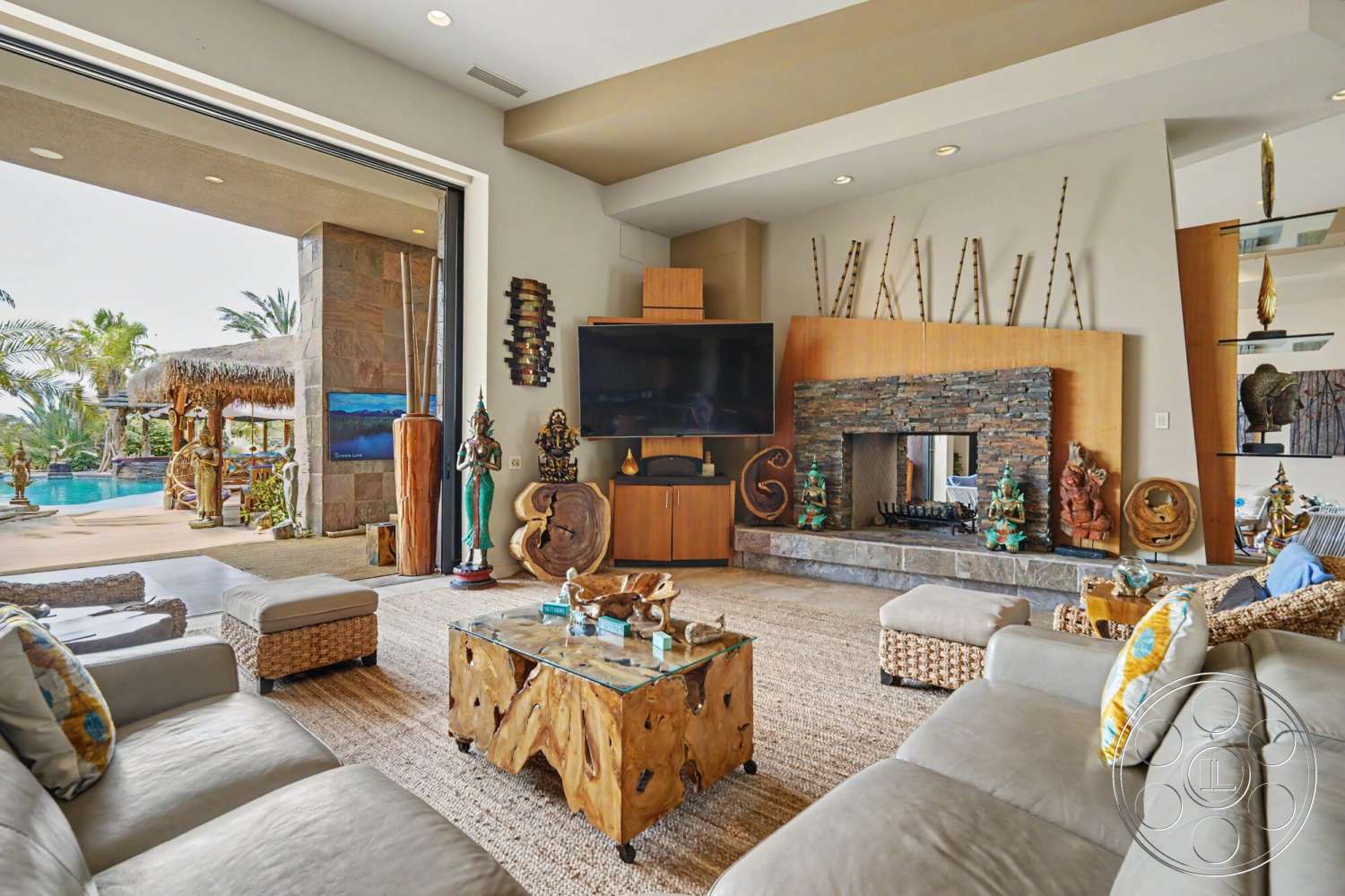 Rancho Mirage 4 - textured area rug, spa-like atmosphere, indoor-outdoor flow, zen-inspired design, palm tree view, stylish home decor, natural wood coffee table, large television, open-concept living area, wood beam ceiling, stone fireplace wall, modern entertainment center, wall-mounted art display, decorative wooden sculptures, folding glass doors, woven rattan armchairs, sectional leather sofa