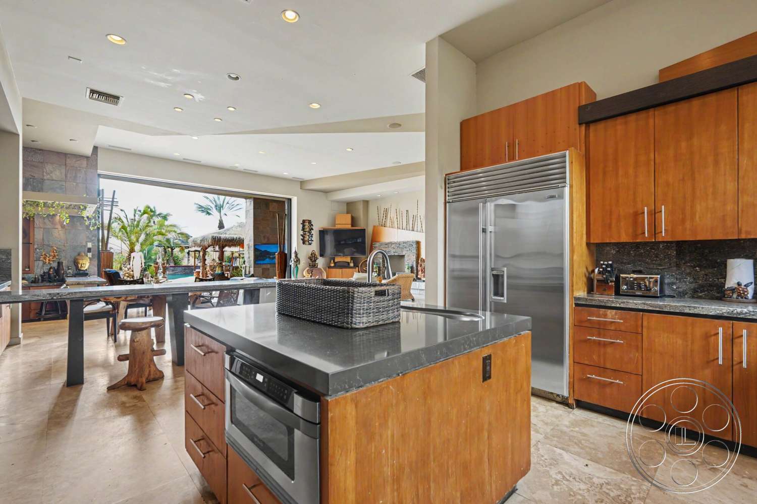 Rancho Mirage 4 - natural light, large glass sliding doors, open floor plan, wood cabinetry, stainless steel refrigerator, light-colored tile flooring, decorative accent pieces, modern kitchen design, granite kitchen countertop, slate stone wall, tropical outdoor view, kitchen island with bar stools