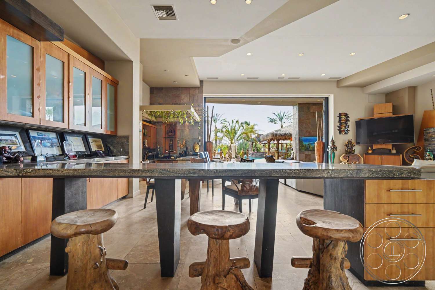 Rancho Mirage 4 - stone accent wall, ceramic tile flooring, modern kitchen design, granite kitchen countertop, exposed ceiling with recessed lighting, contemporary home style, indoor-outdoor living space, open plan interior layout, wood kitchen cabinet with glass doors, natural wood stool seating, lush outdoor palm tree view